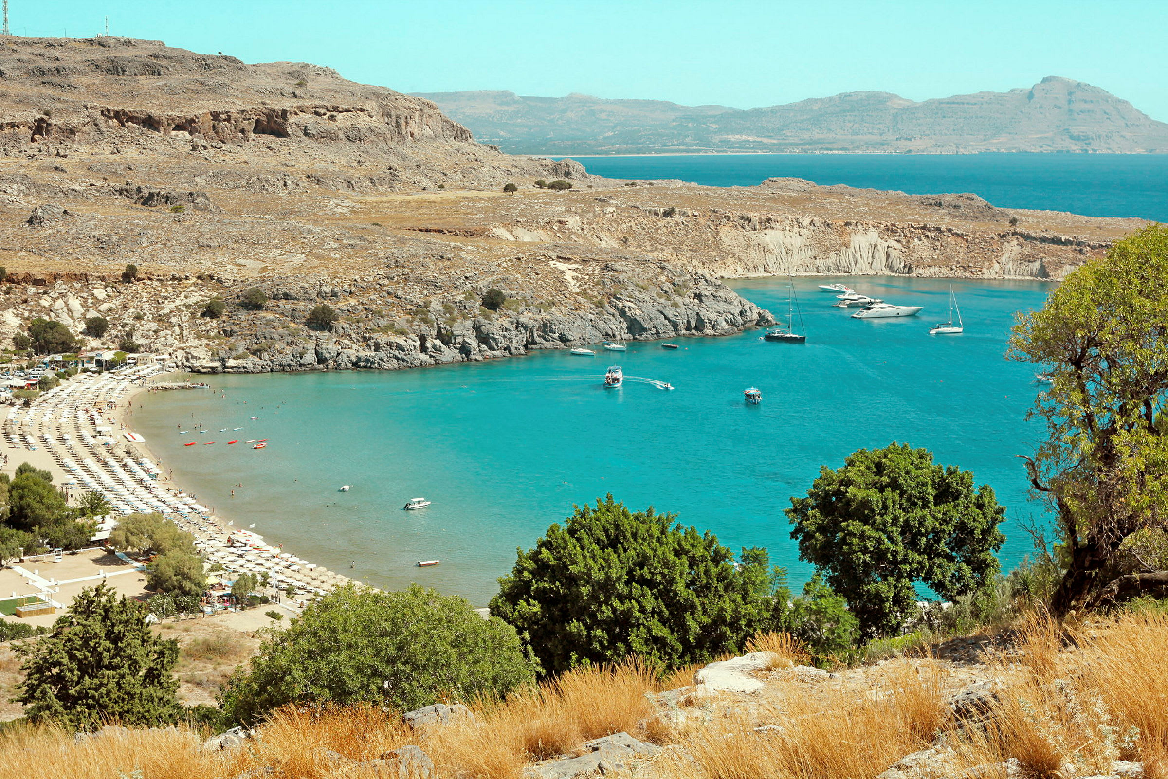 Strand med solsenger og parasoller ved en bukt med turkist vann og båter. (AI-generert tekst)
