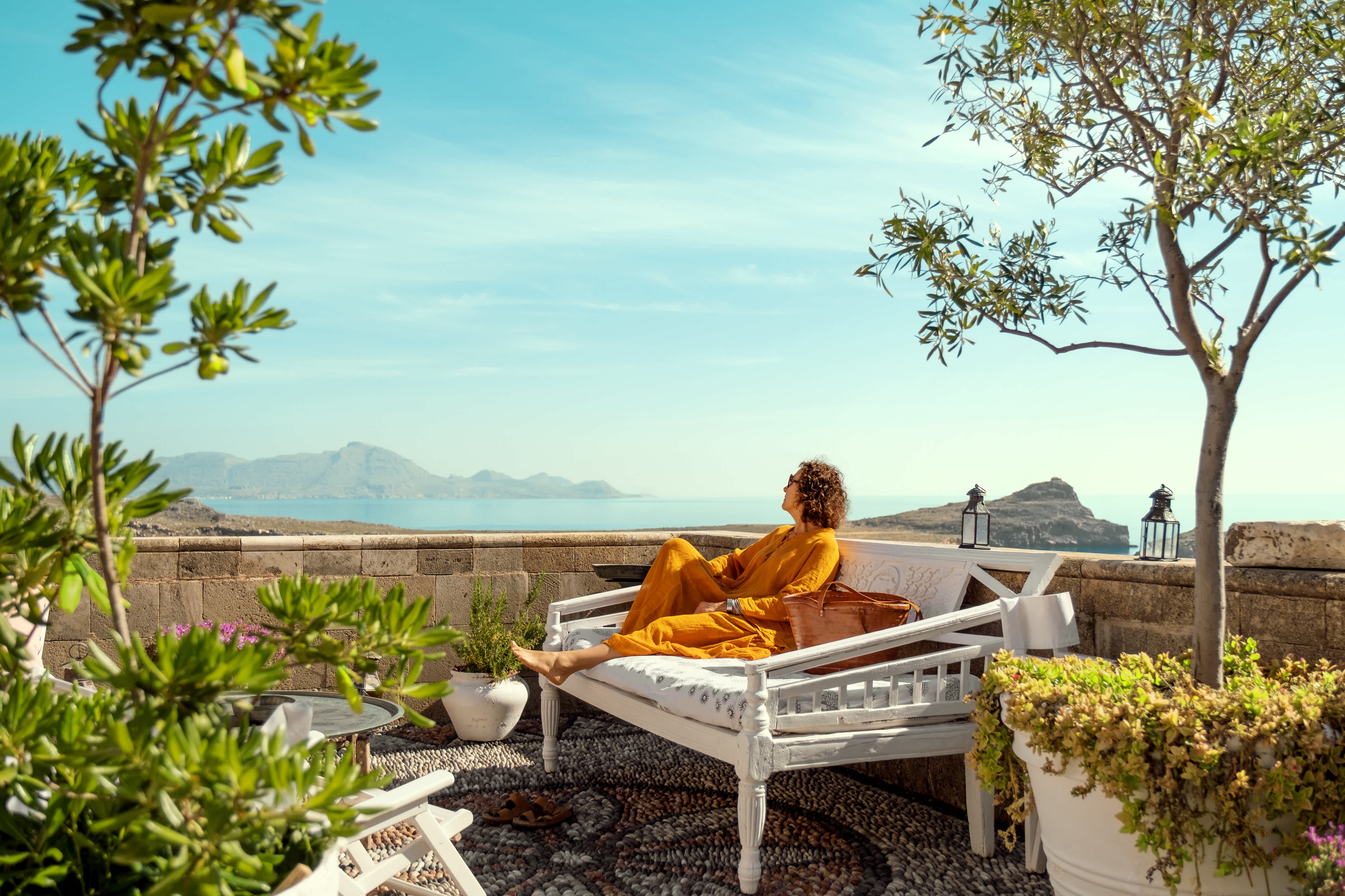 En person slapper av på en hvit benk på en terrasse, omgitt av grønt med utsikt over havet og fjellene. (AI-generert tekst)