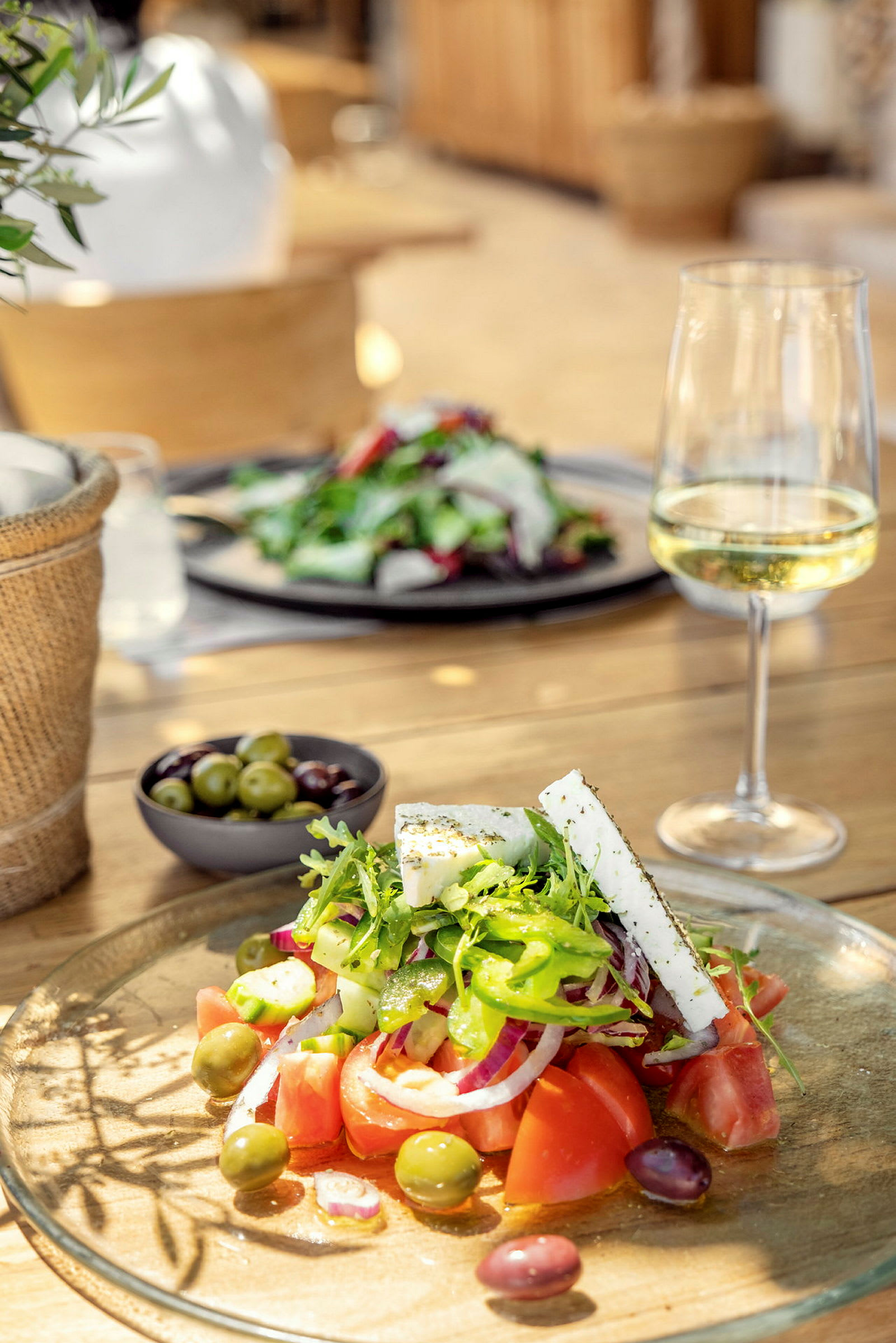 Bilde av en tallerken med salat, fetaost og oliven samt et glass hvitvin på et trebord. (AI-generert tekst)