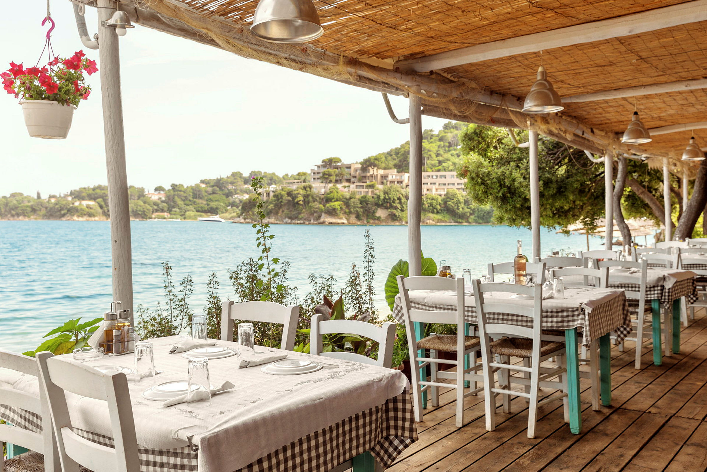 En restaurant med utsikt over havet, med bord som har hvite duker og rutete bordoverlegg. (AI-generert tekst)