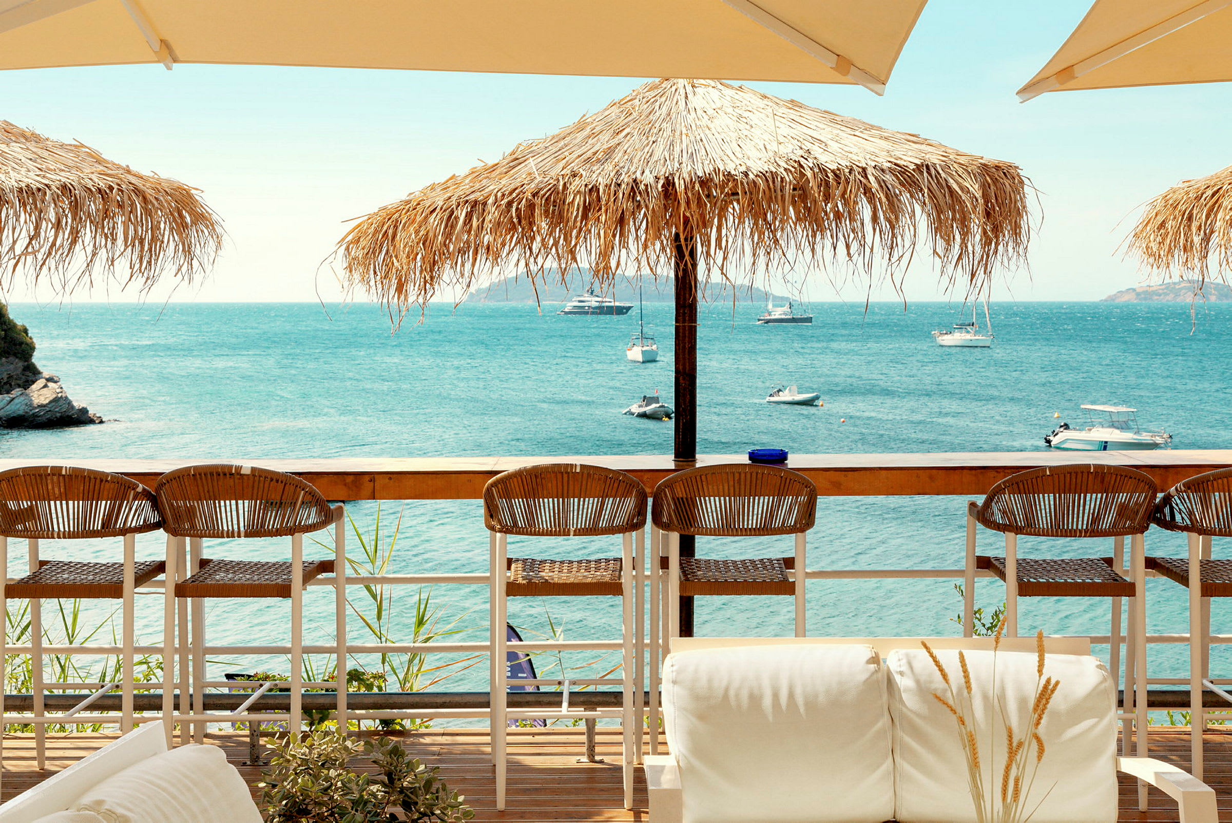 Utsikt over havet med båter, parasoller og sitteplasser ved en strandbar. (AI-generert tekst)
