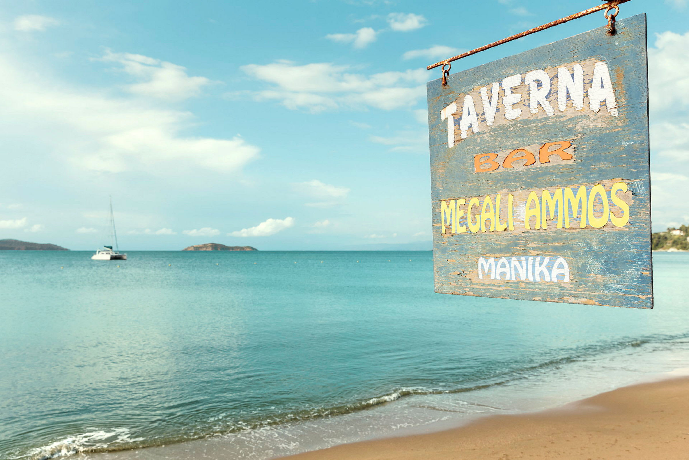 Skilt for en taverna ved en sandstrand med rolig sjø i bakgrunnen. (AI-generert tekst)
