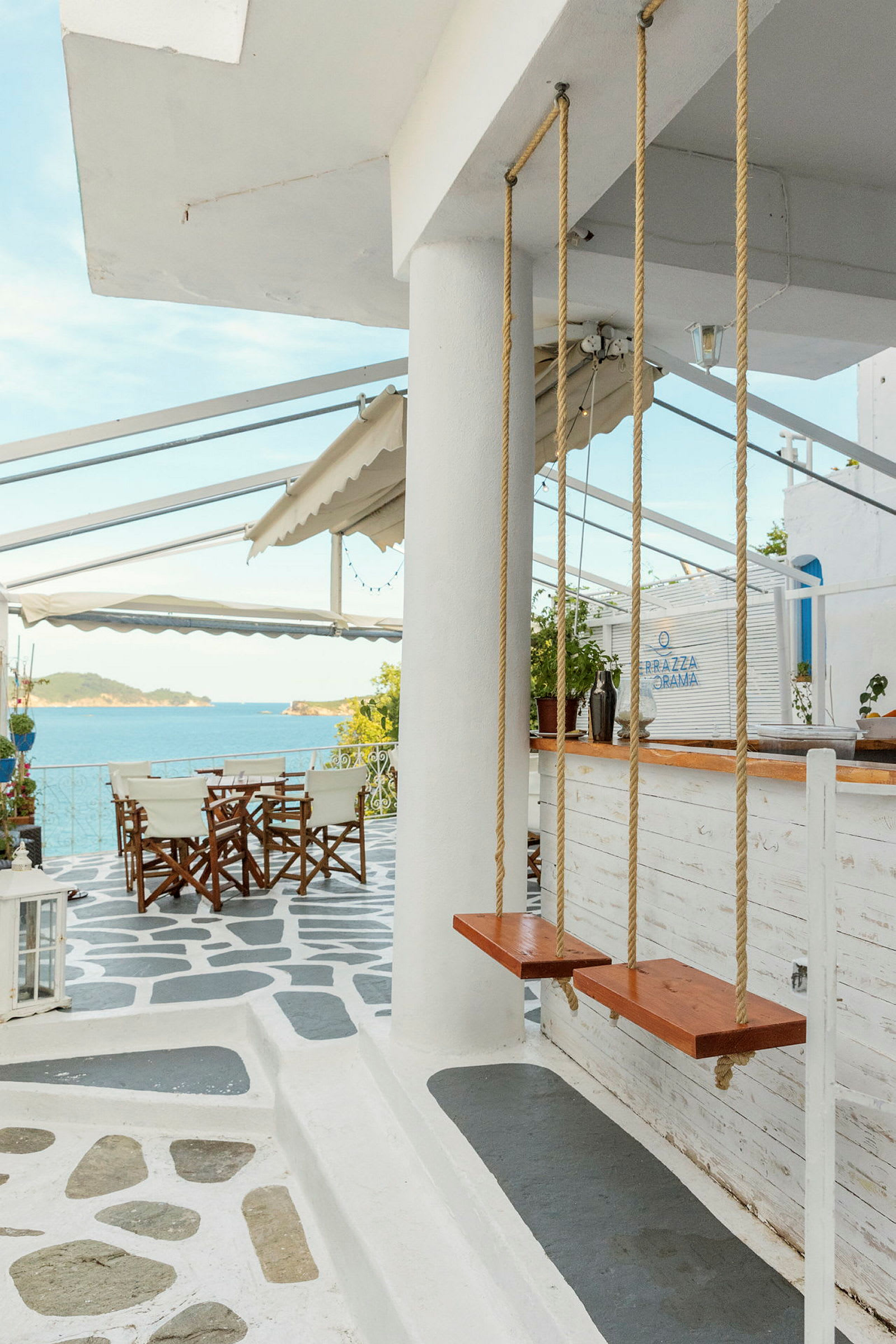Bilde av en terrasse med stoler og bord, havutsikt og to tresvinger foran en søyle. (AI-generert tekst)