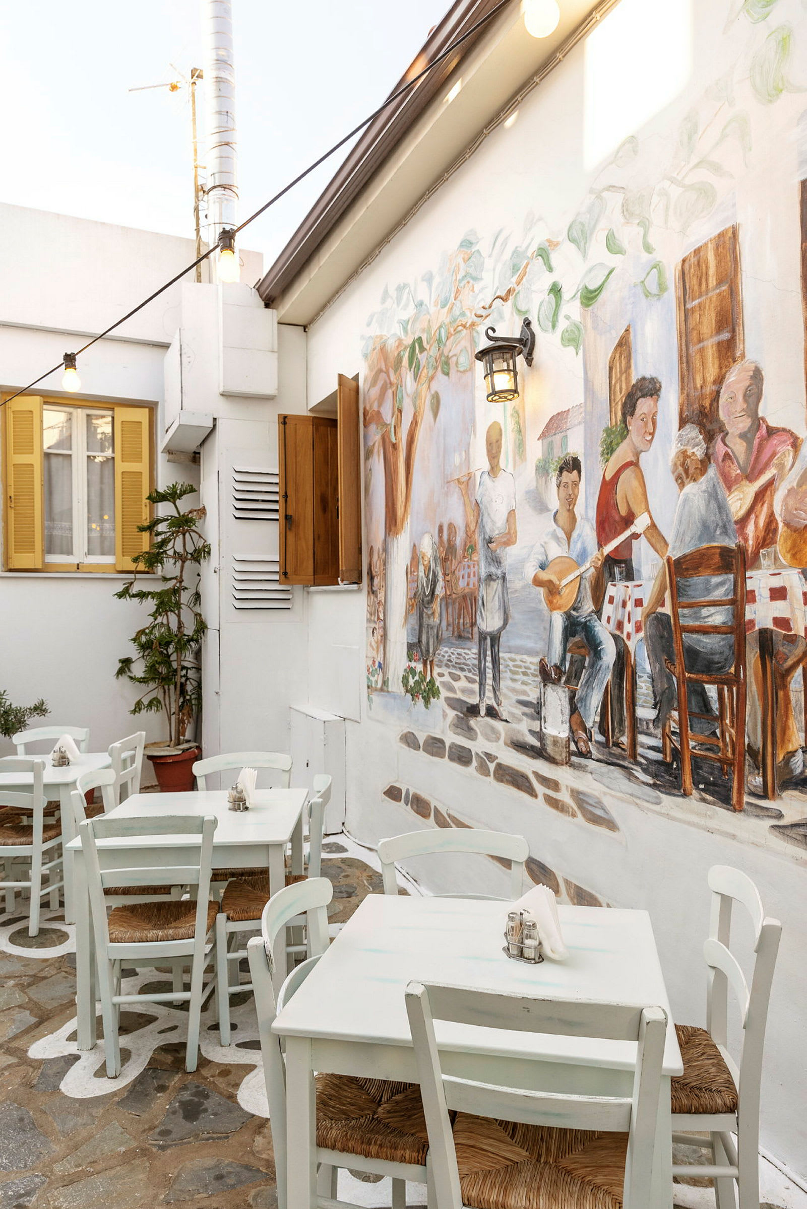 Utendørsområde med hvite bord og stoler, vegg dekorert med maleri av mennesker i en restaurantmiljø. (AI-generert tekst)