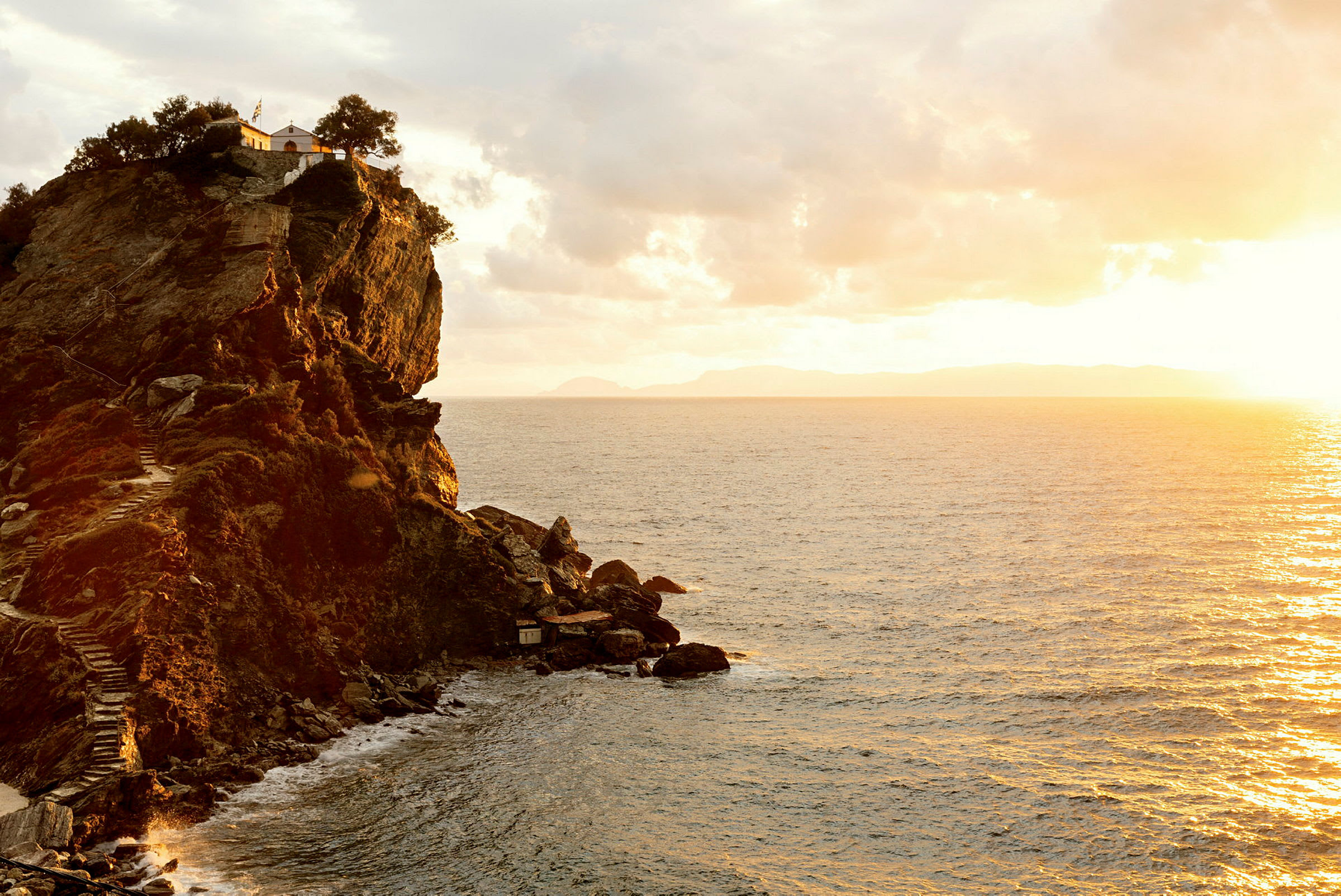 En klippe ved kysten med en bygning og trær på toppen. Havet sees under en solnedgang. (AI-generert tekst)
