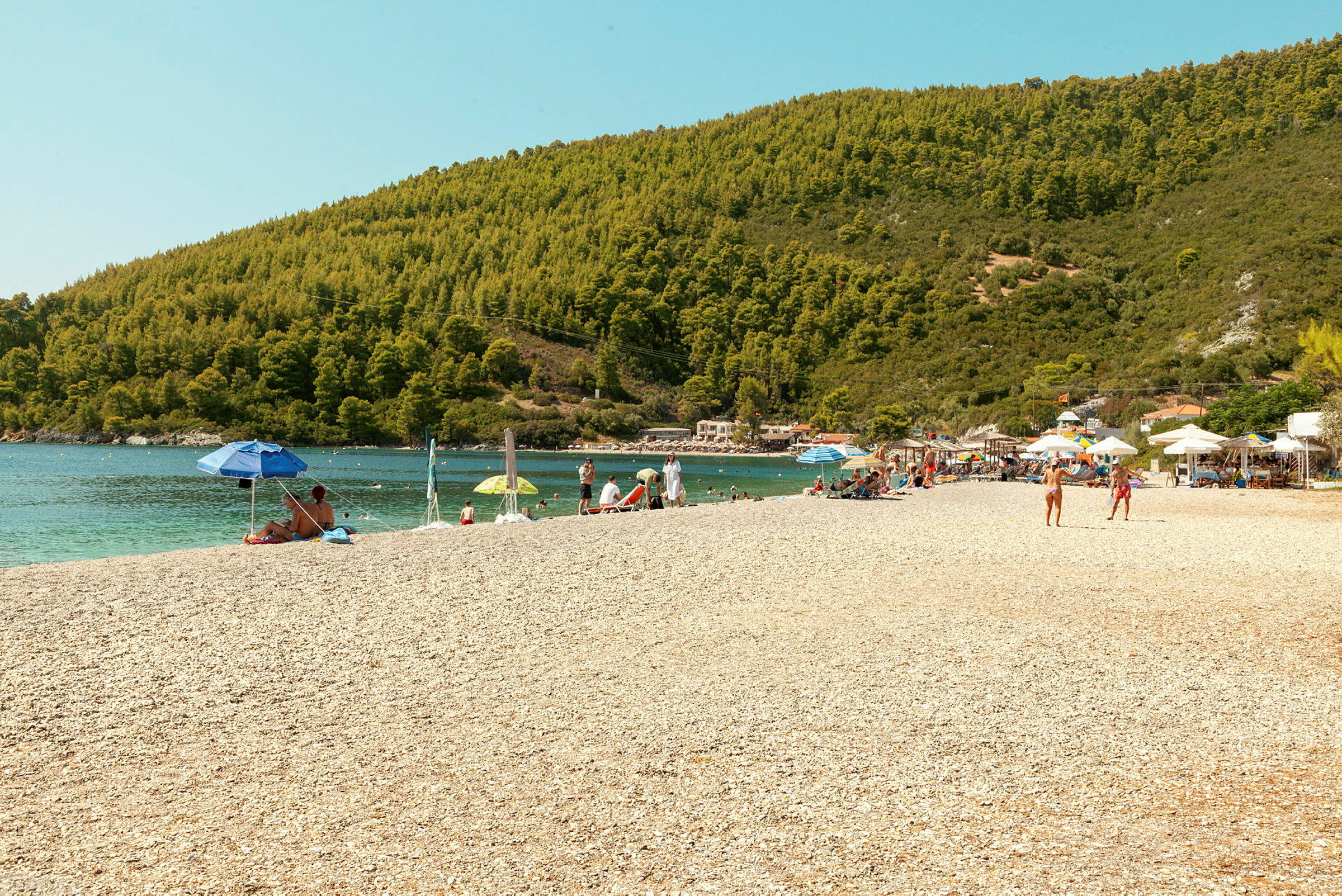En strand med solbadende mennesker, parasoller og omkringliggende grønne åser. (AI-generert tekst)
