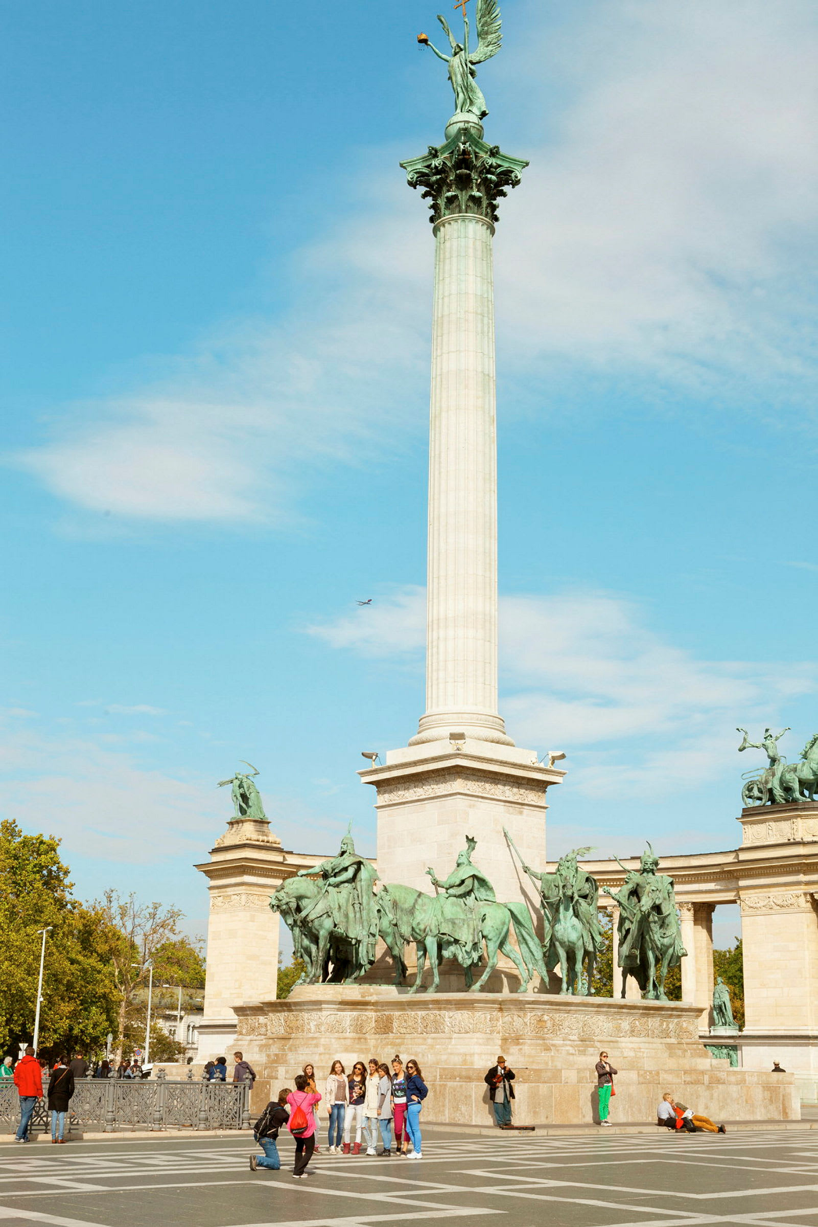 En høy søyle med en statue på toppen, omgitt av flere rytterstatuer ved et monument på en åpen plass. (AI-generert tekst)