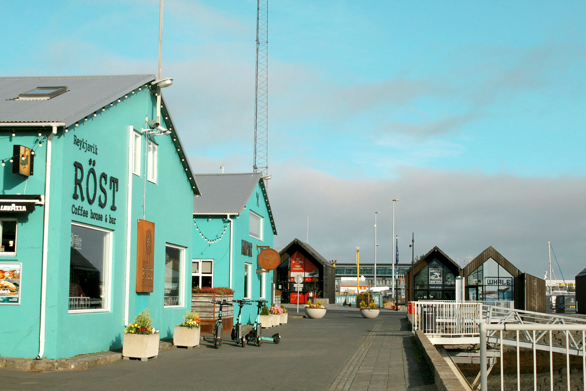 Et turkis kaféhus ved en havn med bygninger og brygge i bakgrunnen. (AI-generert tekst)
