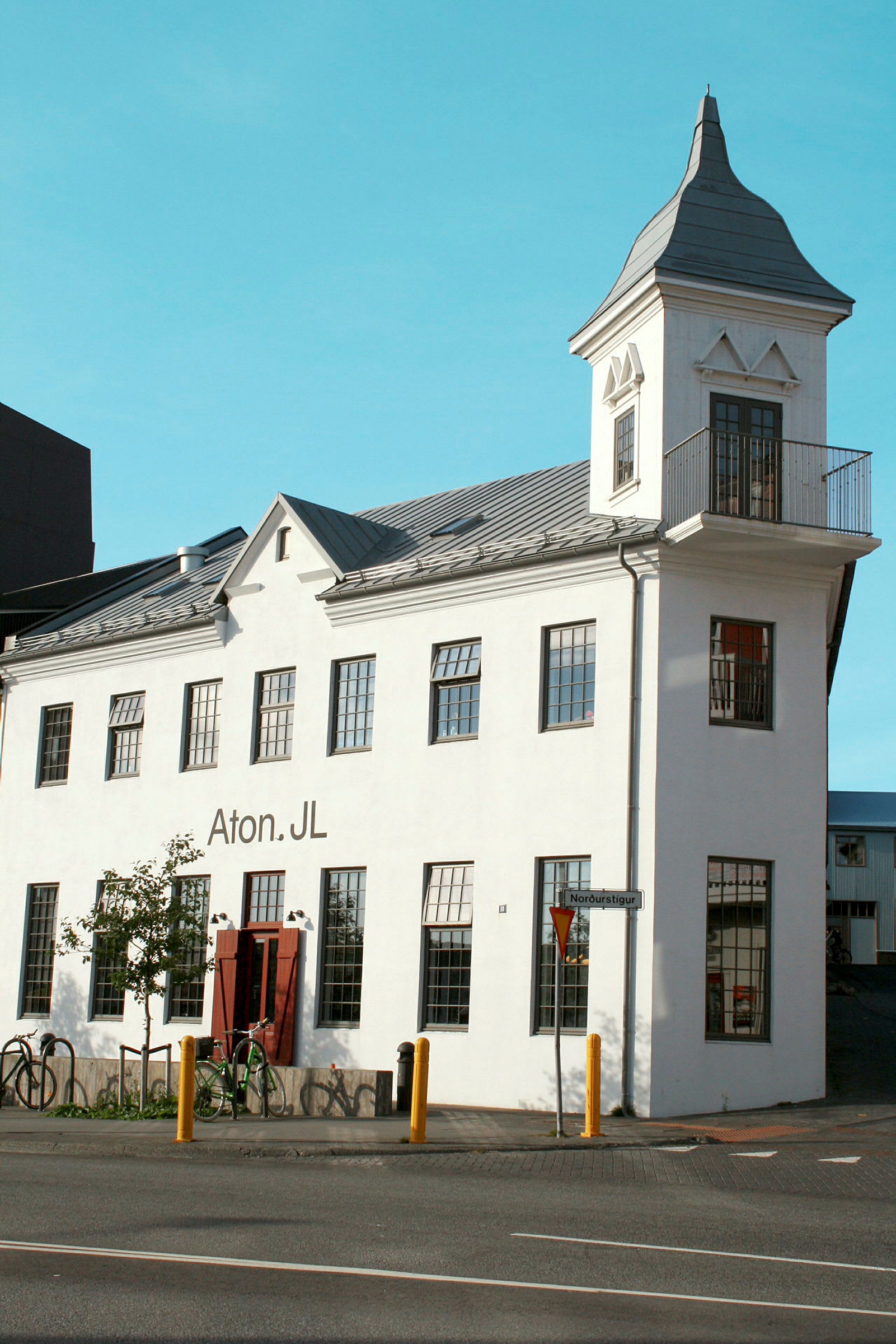 En hvit bygning med et tårn og store vinduer. Bygningen har et skilt som sier "Aton.JL", og det står sykler parkert foran. (AI-generert tekst)
