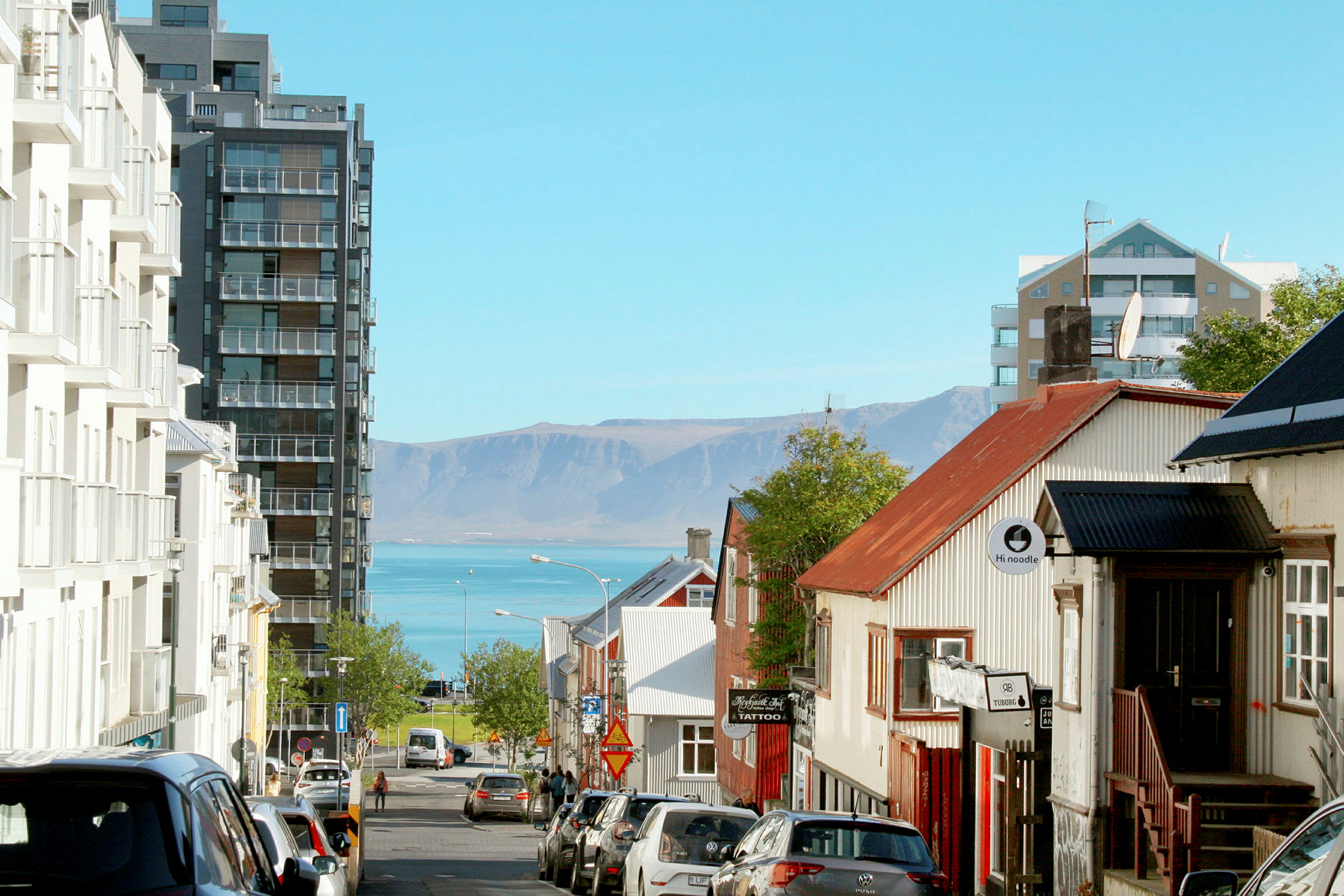 En gate i en by med bygninger på begge sider og utsikt over havet og fjell i bakgrunnen. (AI-generert tekst)