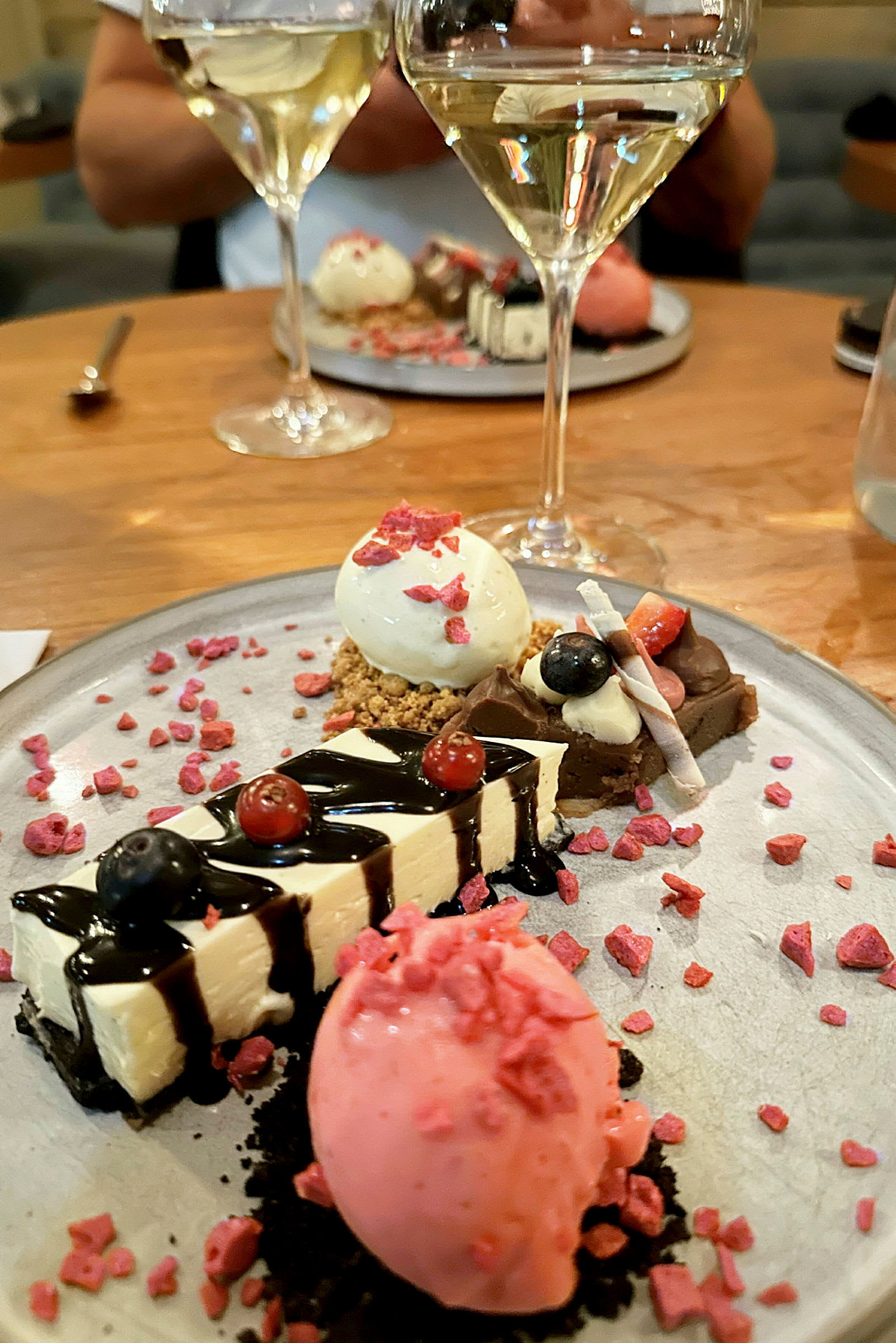 En tallerken med ulike desserter, inkludert en rosa sorbet, ostekake og andre små søtsaker, dekorert med bær og saus. To glass hvitvin kan sees i bakgrunnen. (AI-generert tekst)