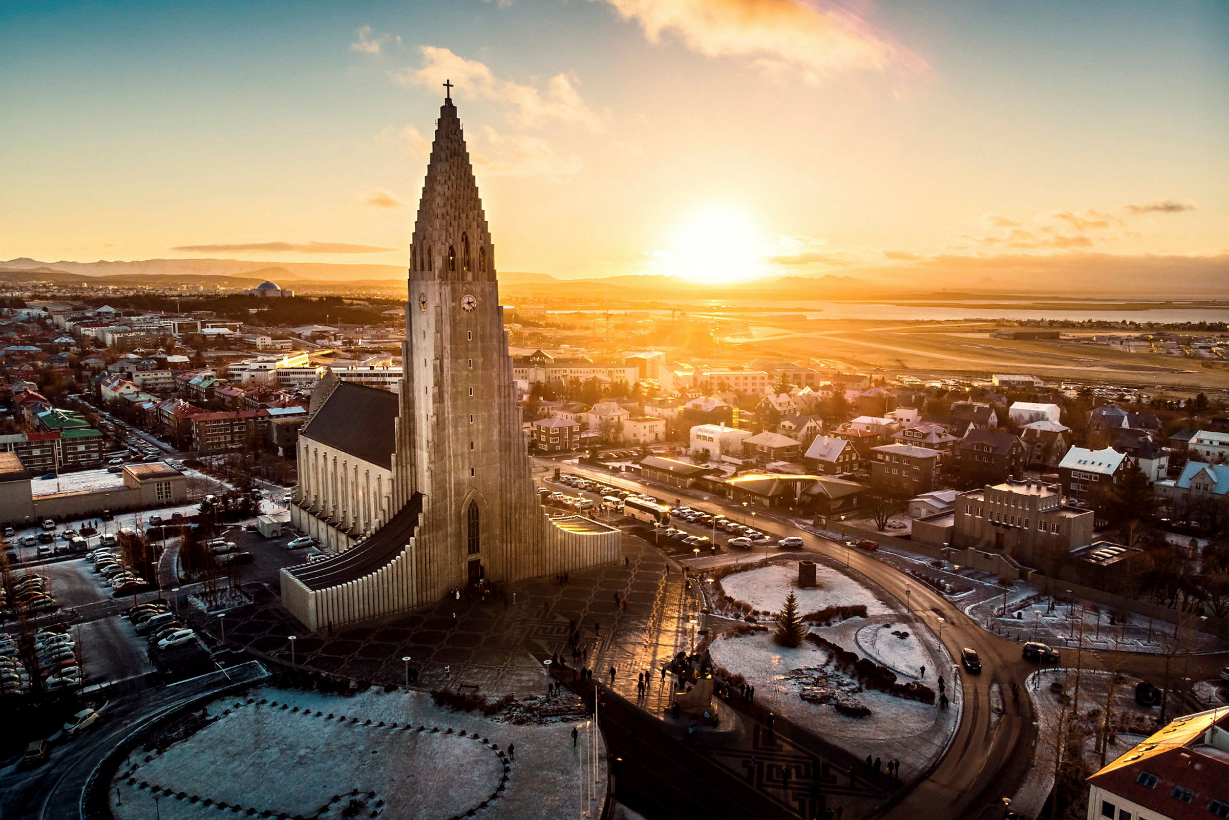 En kirke med moderne arkitektur og en høy tårnstruktur, omgitt av en by og et landskap under en solnedgang. (AI-generert tekst)