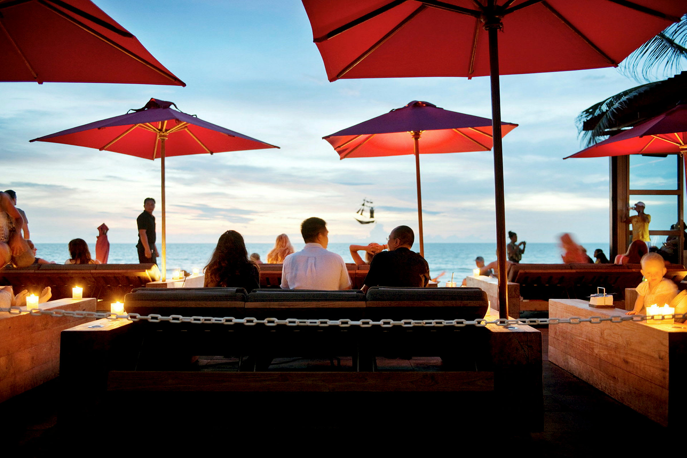 Folk sitter ved et bord på en utestaurant med røde parasoller og havet i bakgrunnen. (AI-generert tekst)