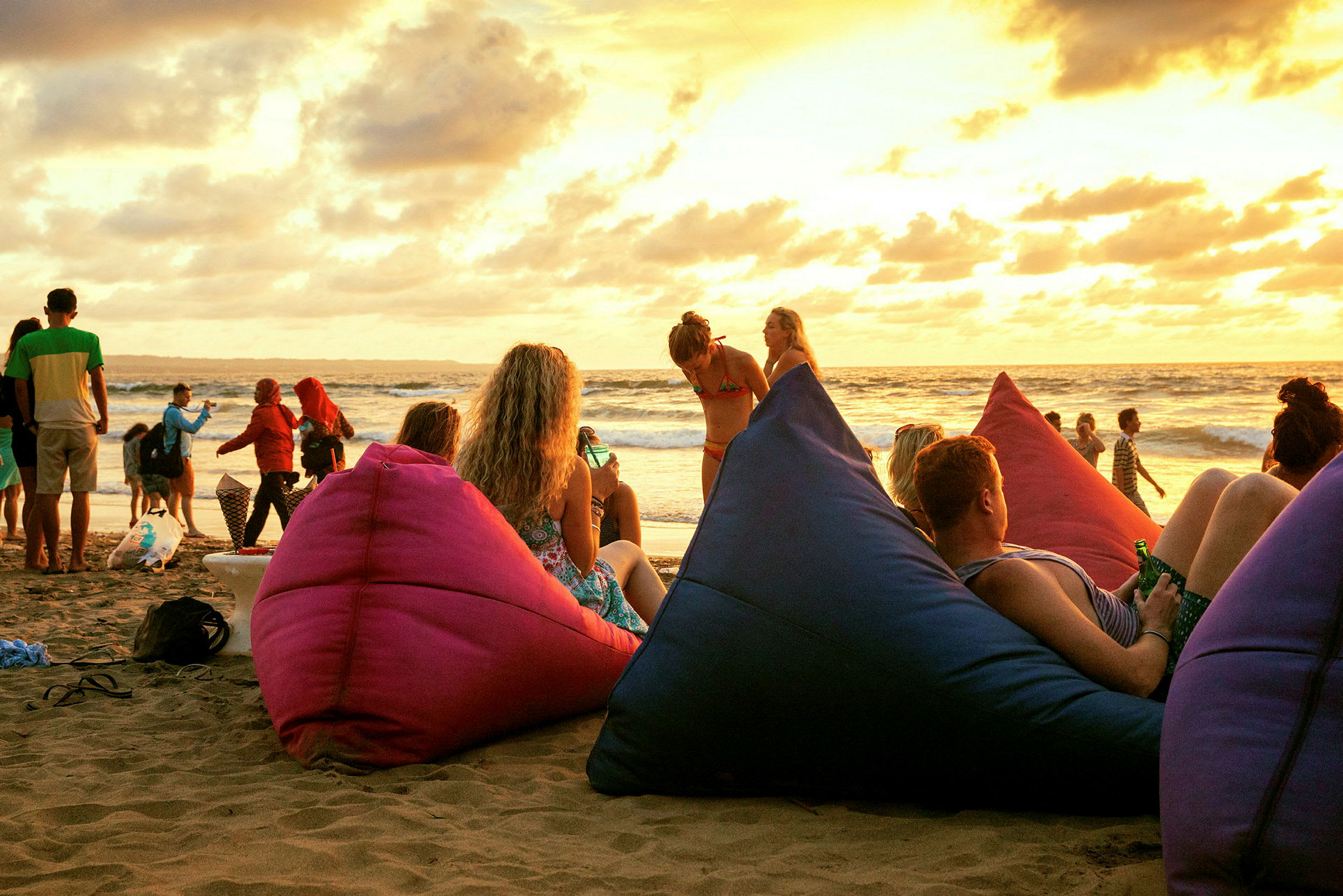 Avslappet strandscene ved solnedgang med mennesker som sitter på fargerike sittesekker. (AI-generert tekst)