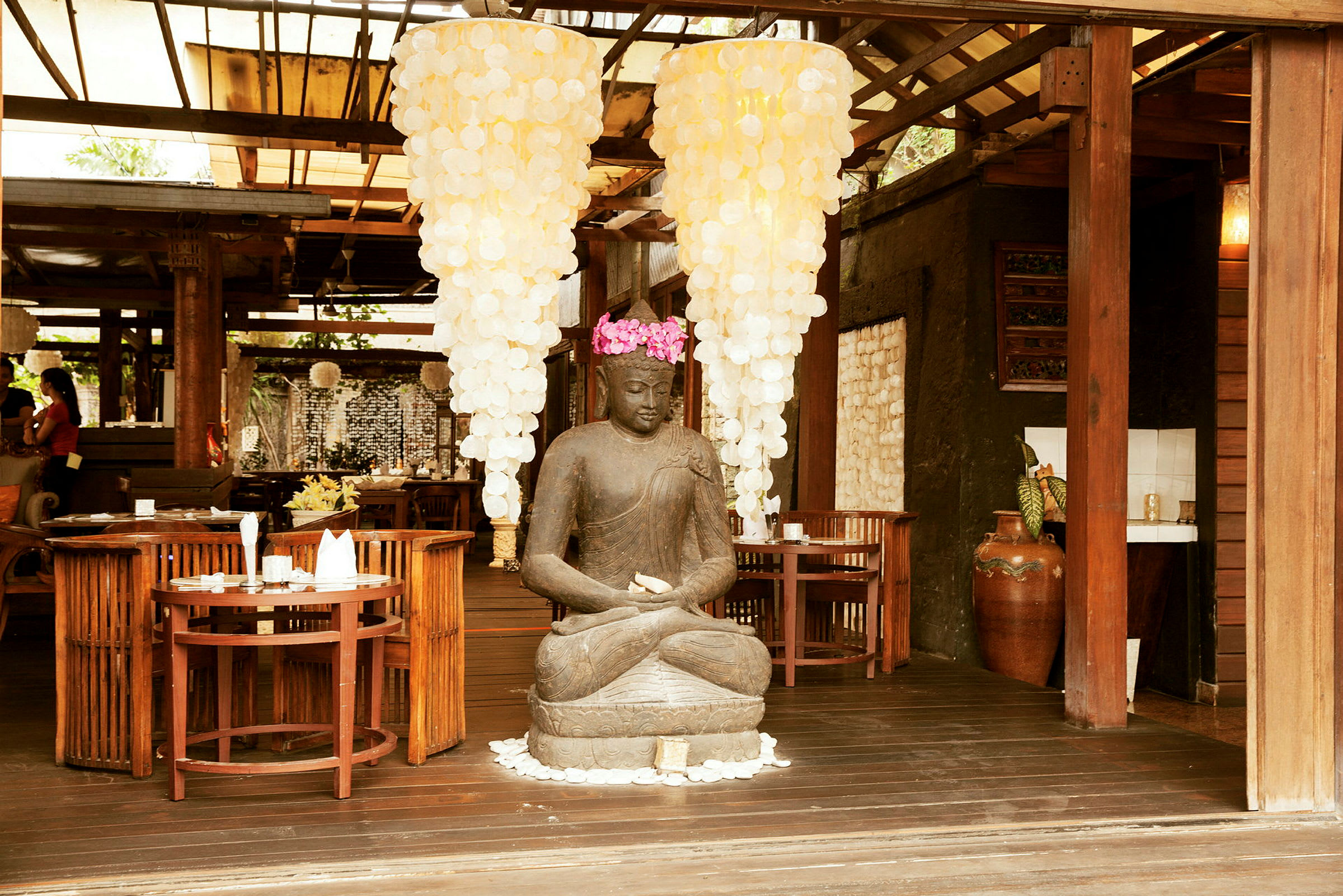 Statue av sittende Buddha omgitt av trebord og stoler, med store lysekroner hengende ovenfor. (AI-generert tekst)