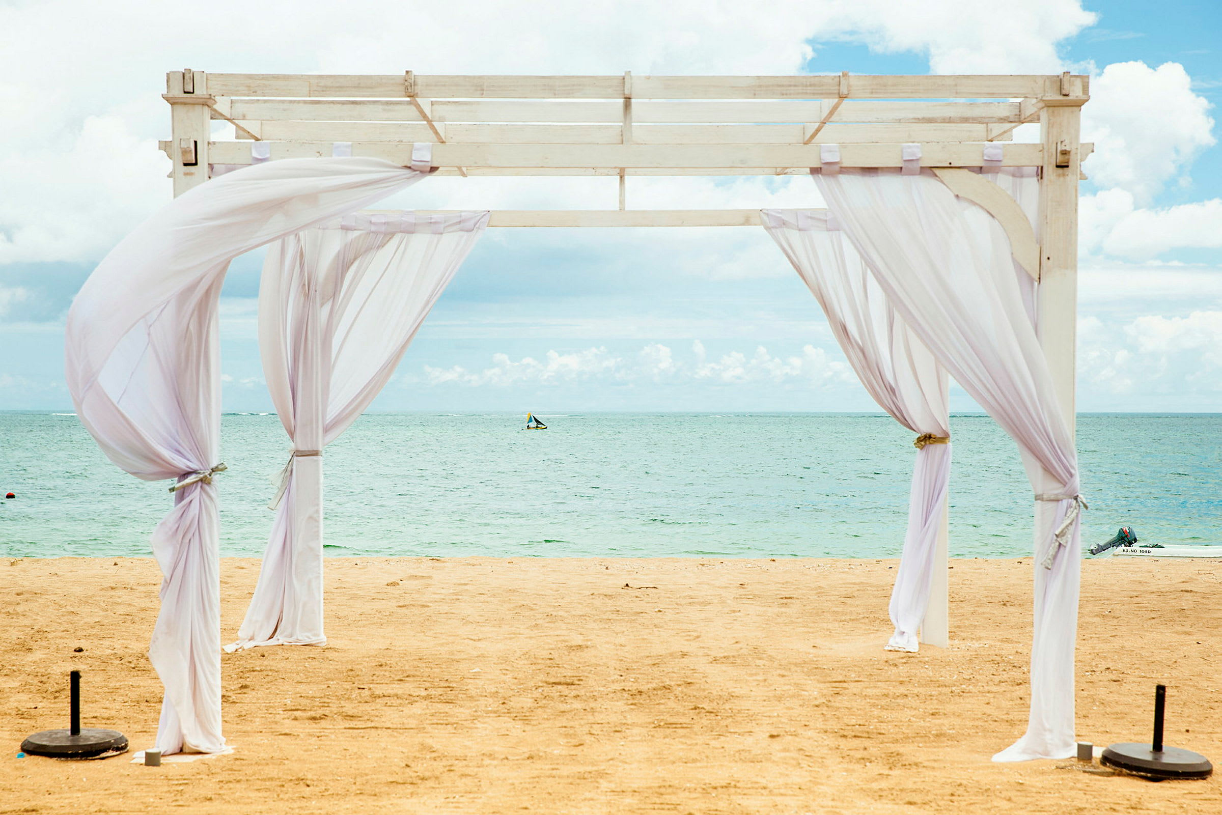 Bildet viser en hvit pergola med tynne stoffer på en sandstrand ved havet. (AI-generert tekst)