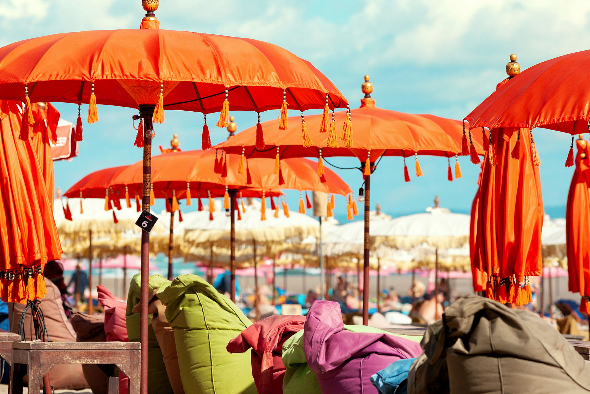 Flere oransje parasoller på rad med fargerike sitteputer under mot en strandbakgrunn. (AI-generert tekst)