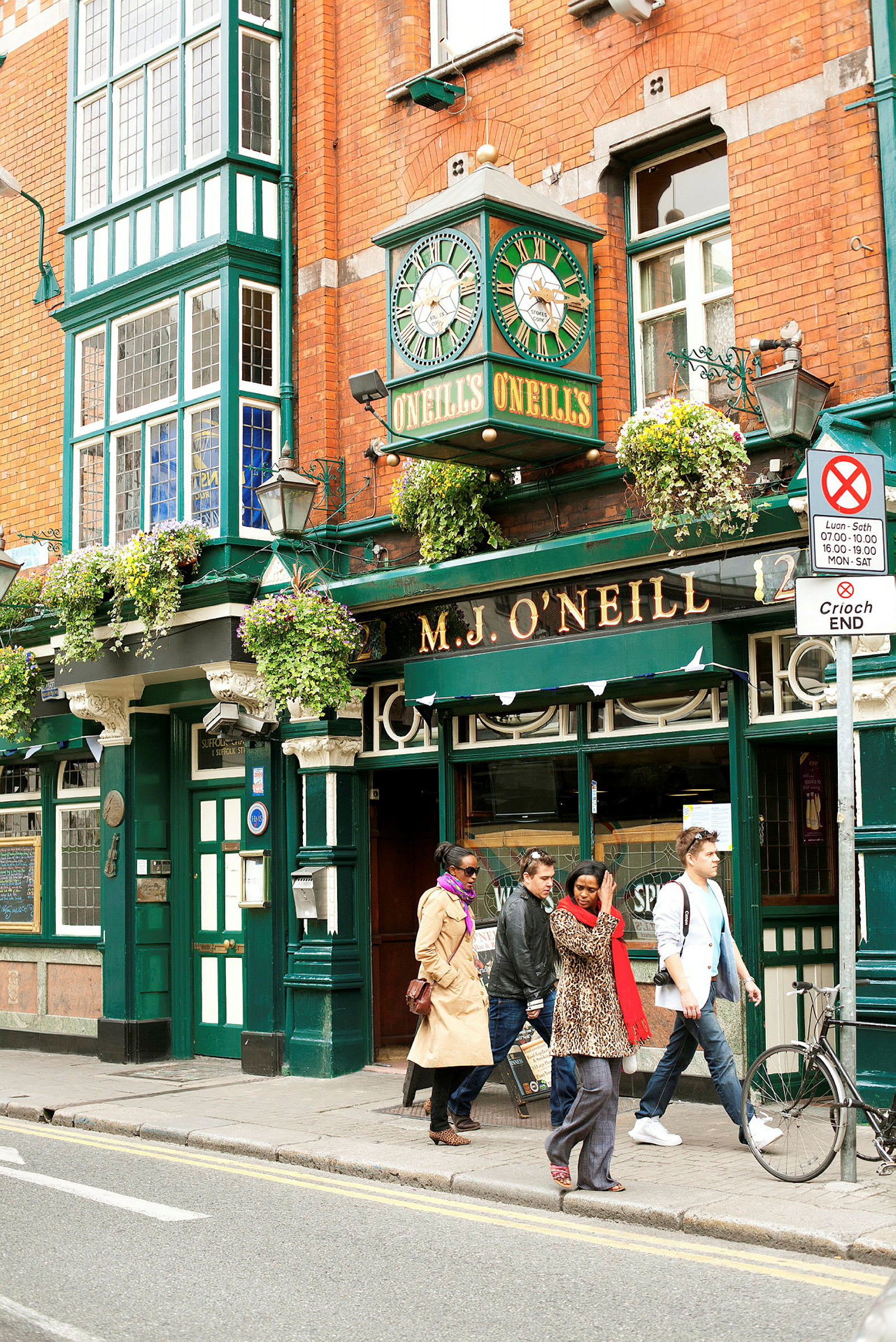 En tradisjonell irsk pub med murvegger og et grønt klokketårn over inngangen. Flere personer går forbi utenfor. (AI-generert tekst)