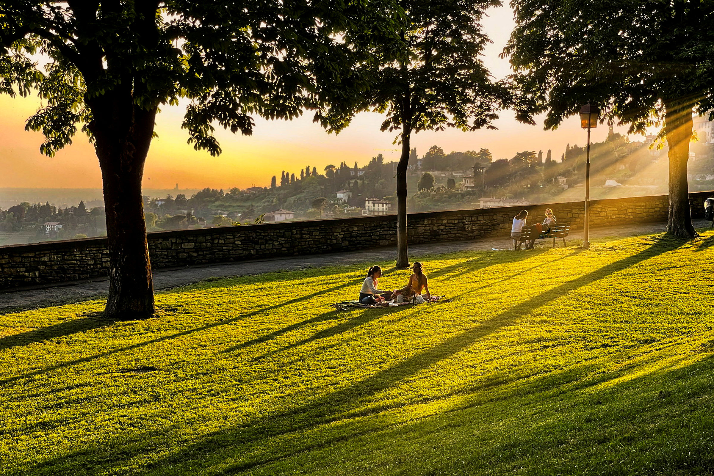 En gressplen ved solnedgangen der folk sitter på tepper og benker under trærnes skygge. (AI-generert tekst)