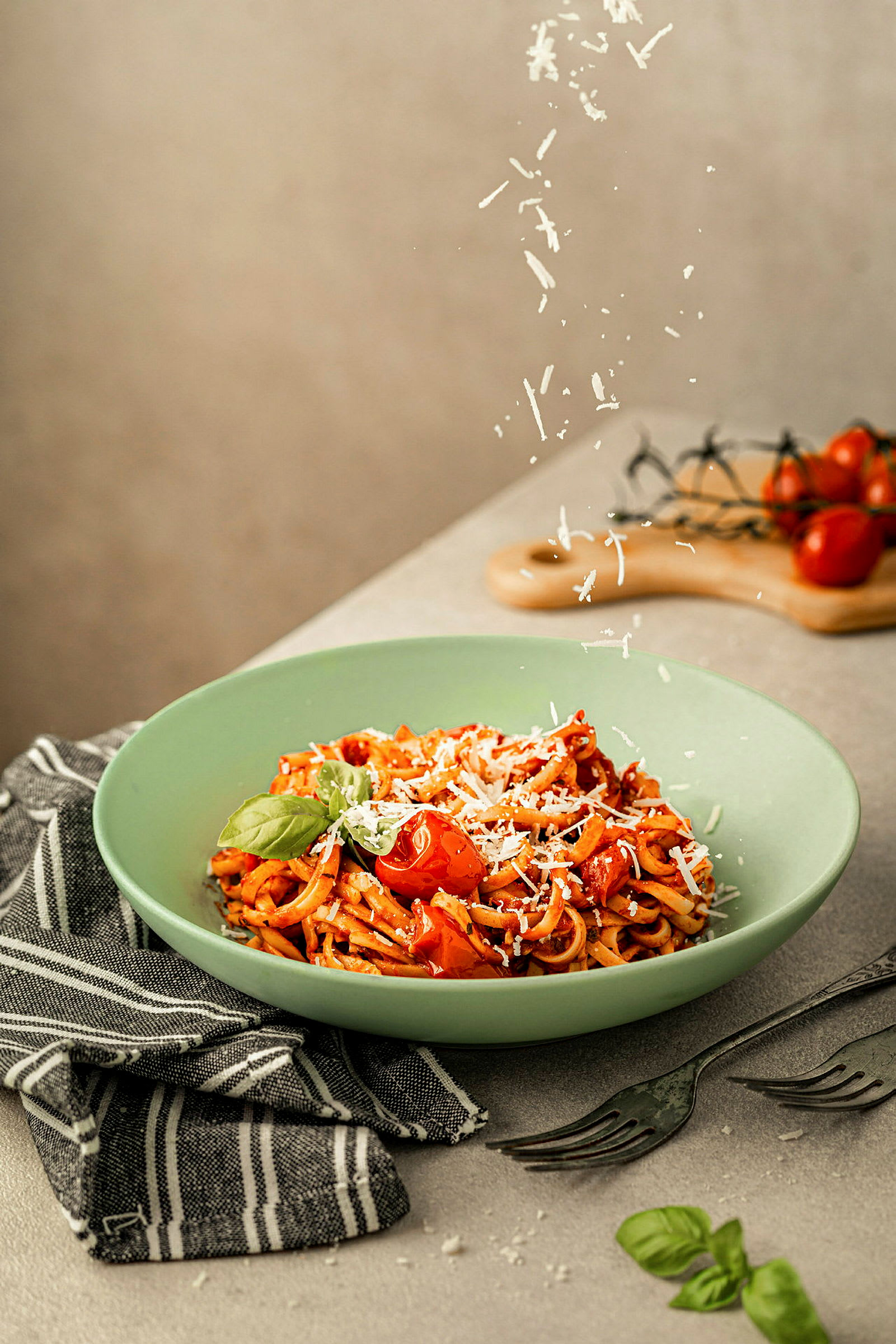 En tallerken med pasta, toppet med tomater, basilikum og revet ost, plassert på en grå overflate med bestikk og et svart-hvitt tøy. (AI-generert tekst)