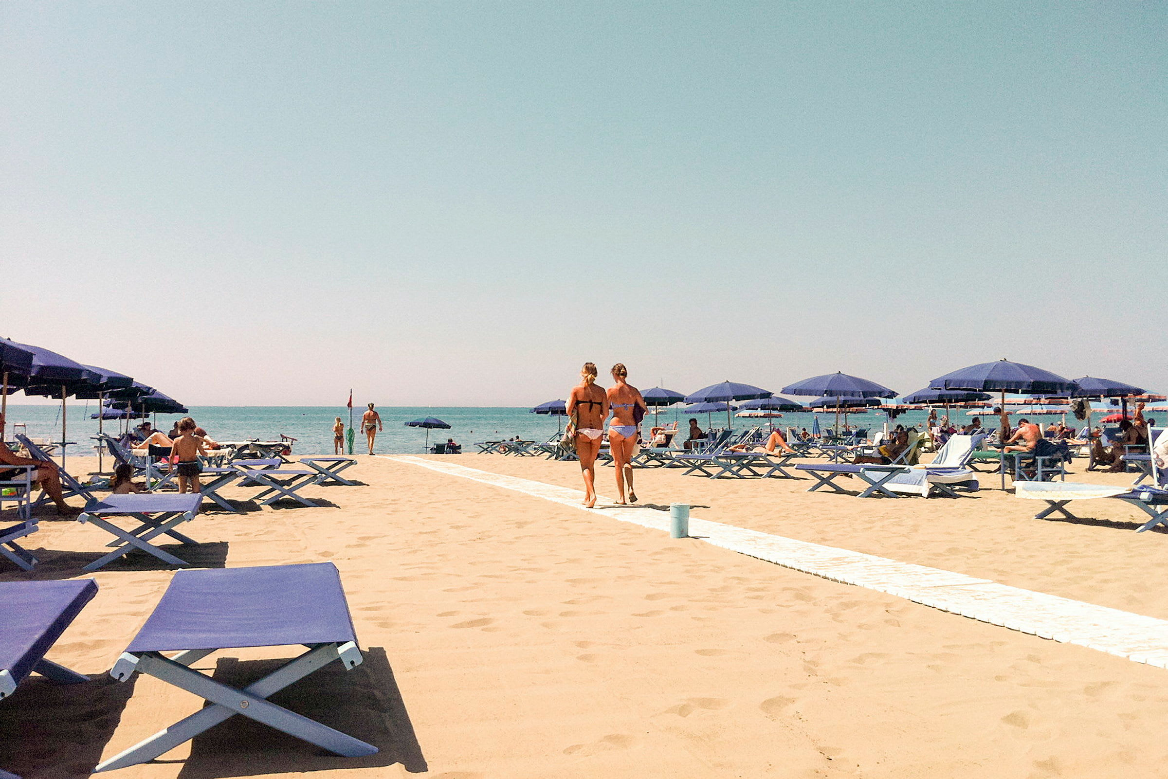 En sandstrand med blå solsenger og parasoller. Folk soler seg og går på stranden med en klar blå himmel i bakgrunnen. (AI-generert tekst)