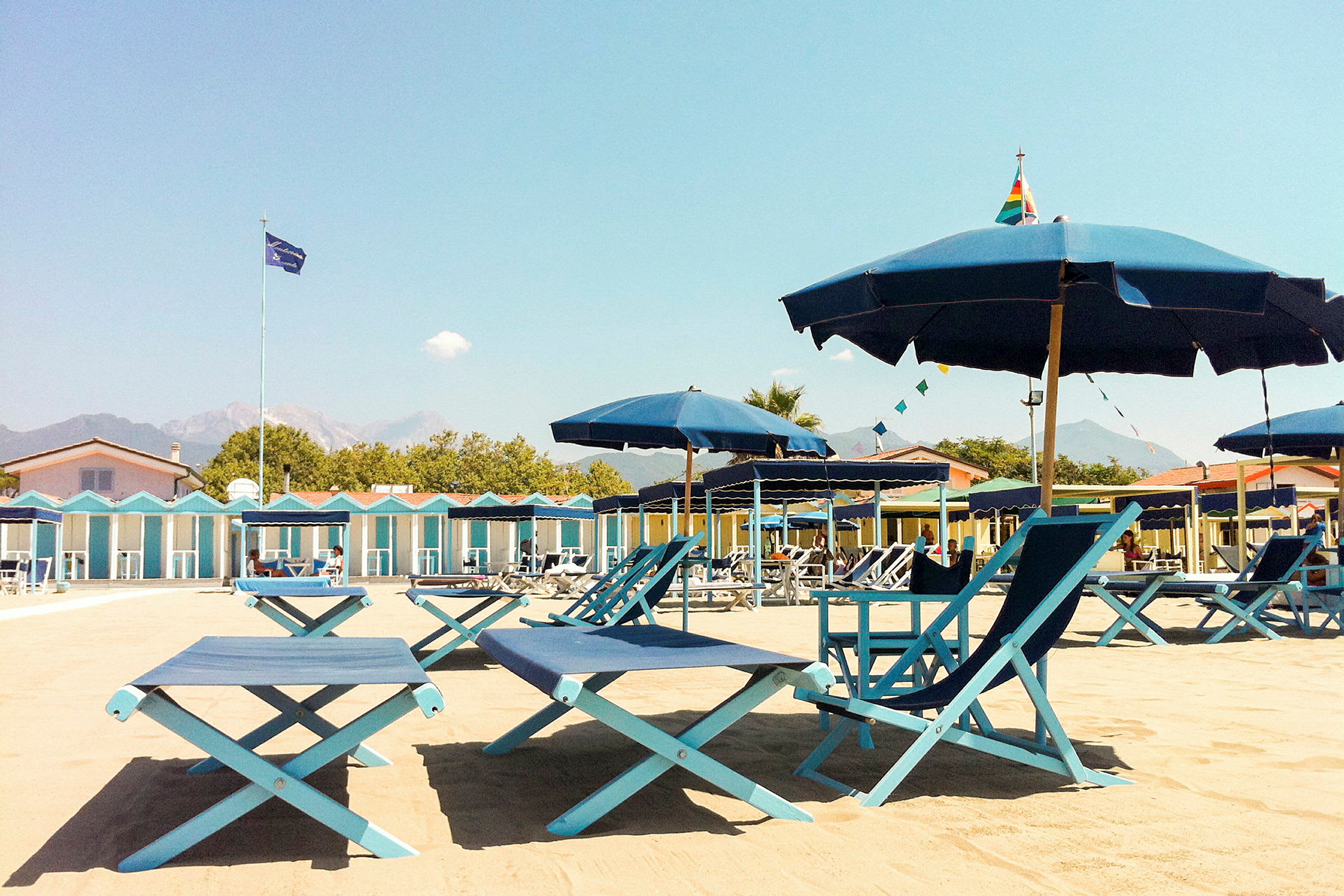 Strandmiljø med solsenger og parasoller samt et område med strandhytter i bakgrunnen. (AI-generert tekst)