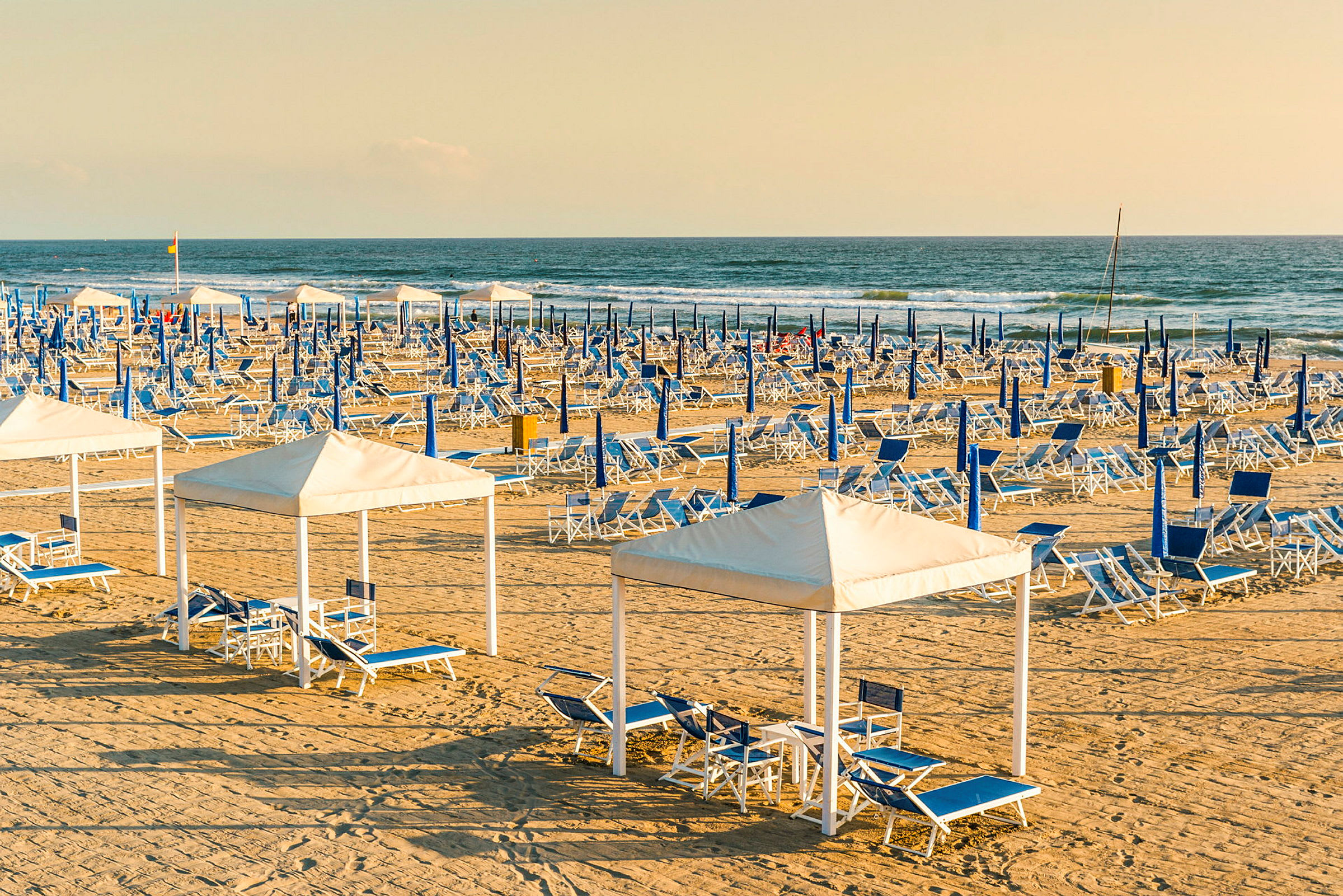 En sandstrand med oppstilte blå og hvite solsenger samt parasoller. I forgrunnen er det cabanas med sittegrupper. (AI-generert tekst)