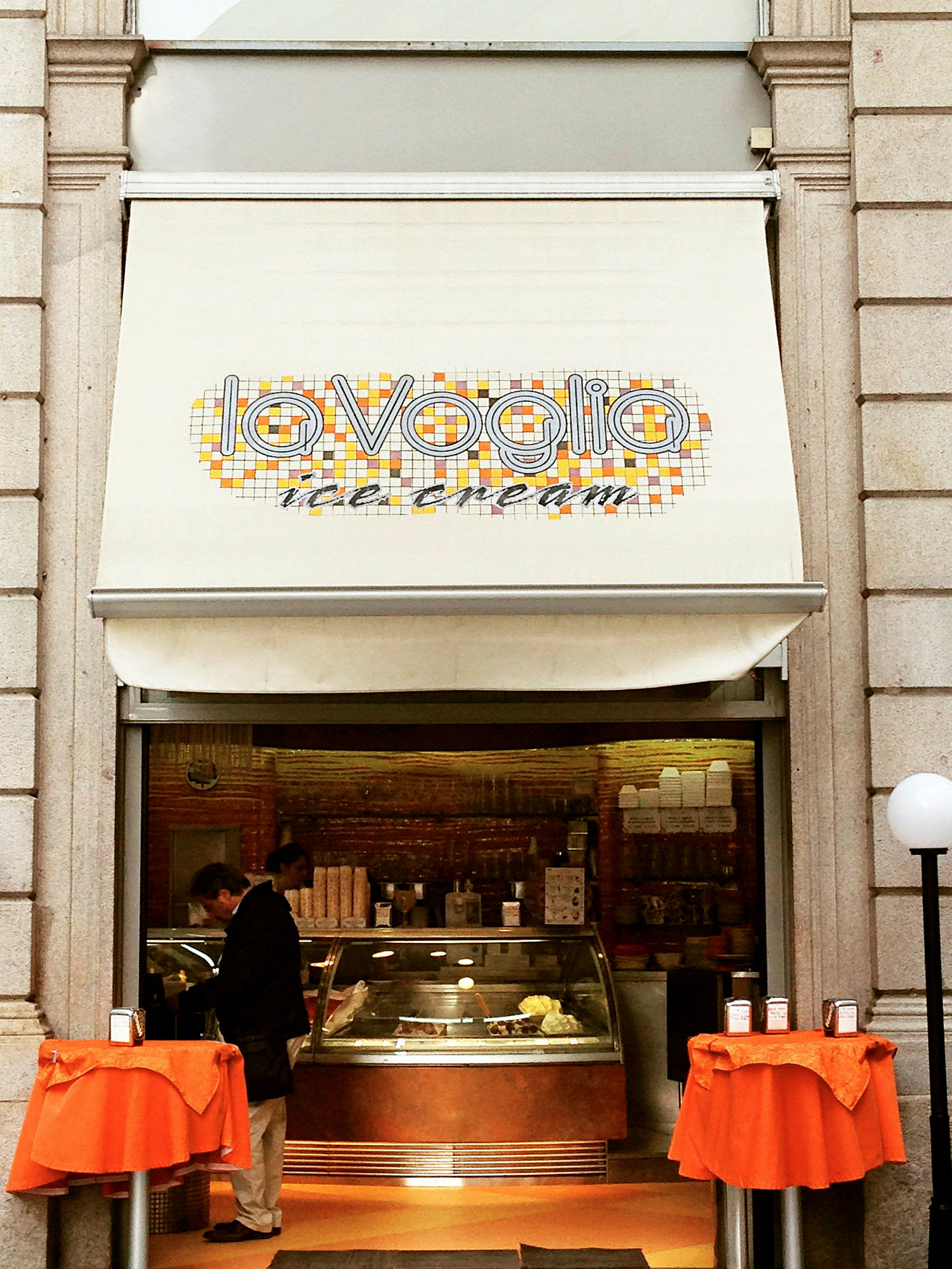 En iskrembutikk med skilt som sier "La Voglia ice cream" og to små bord med oransje duker foran inngangen. (AI-generert tekst)