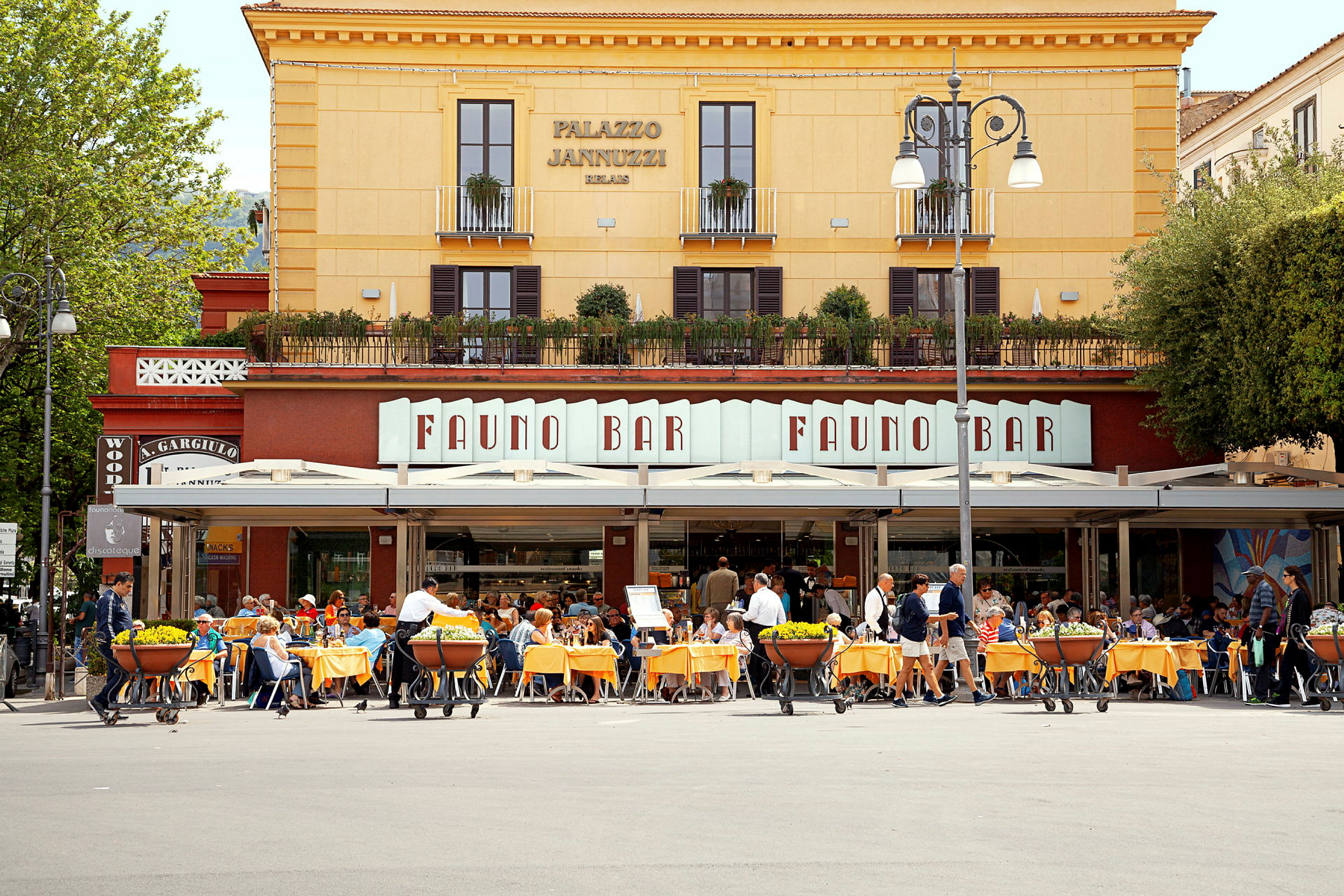 En restaurant med uteservering foran en gul bygning med skilt som viser navnet 'Fauno Bar'. (AI-generert tekst)