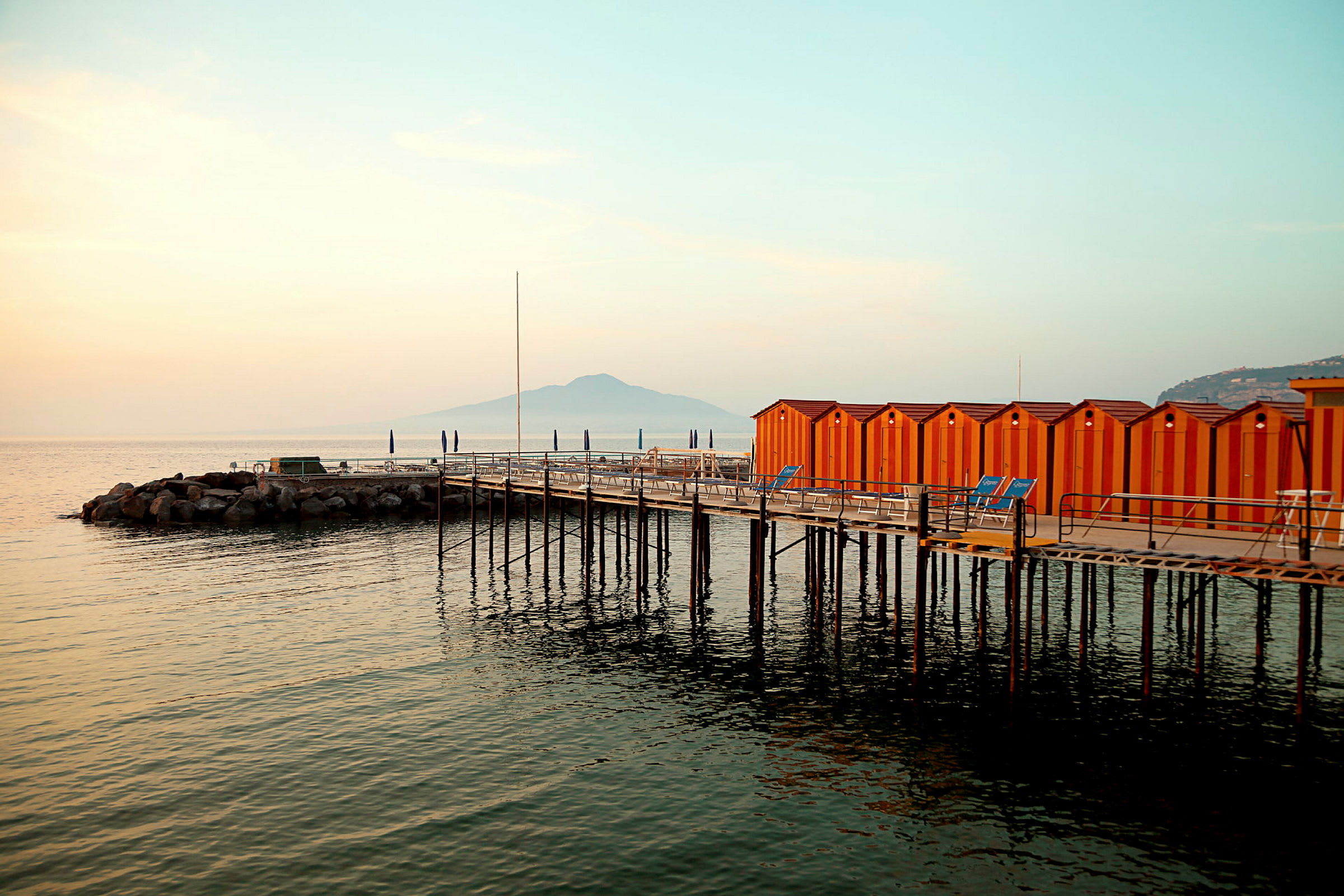 En brygge med oransje hytter ved havet, fjell vises i bakgrunnen. (AI-generert tekst)