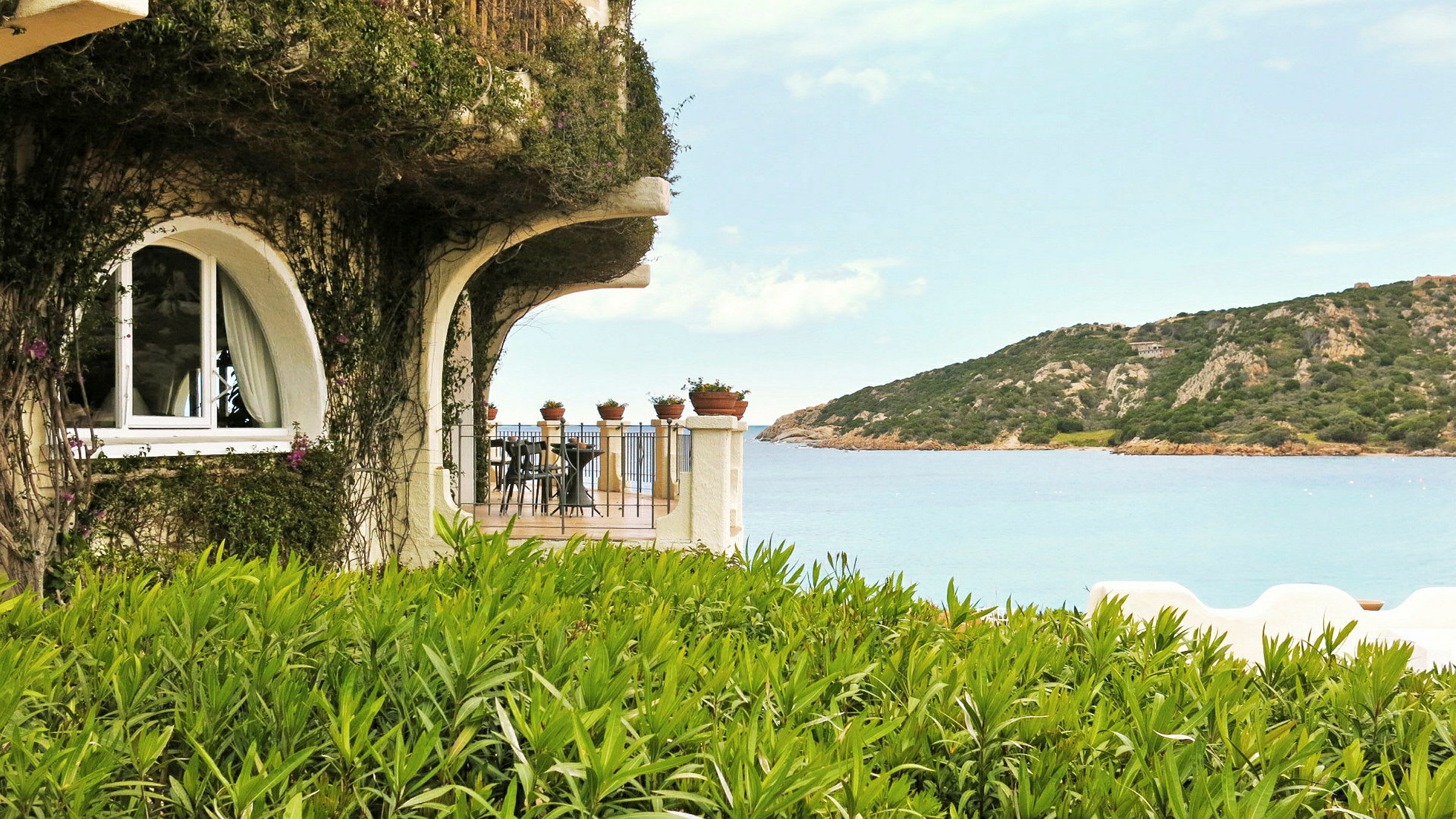 Et hus med grønne planter på fasaden, en terrasse med bord og stoler, samt utsikt over havet og en ås i bakgrunnen. (AI-generert tekst)
