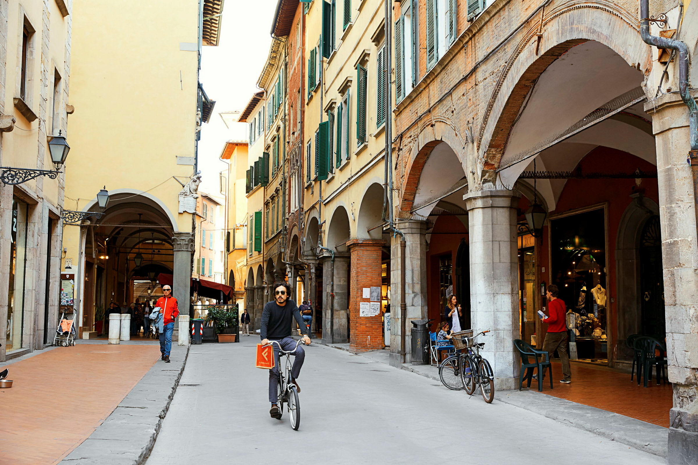 Gate med historiske bygninger og arkader. En person sykler forbi, og noen personer spaserer eller sitter ved en kafé. (AI-generert tekst)
