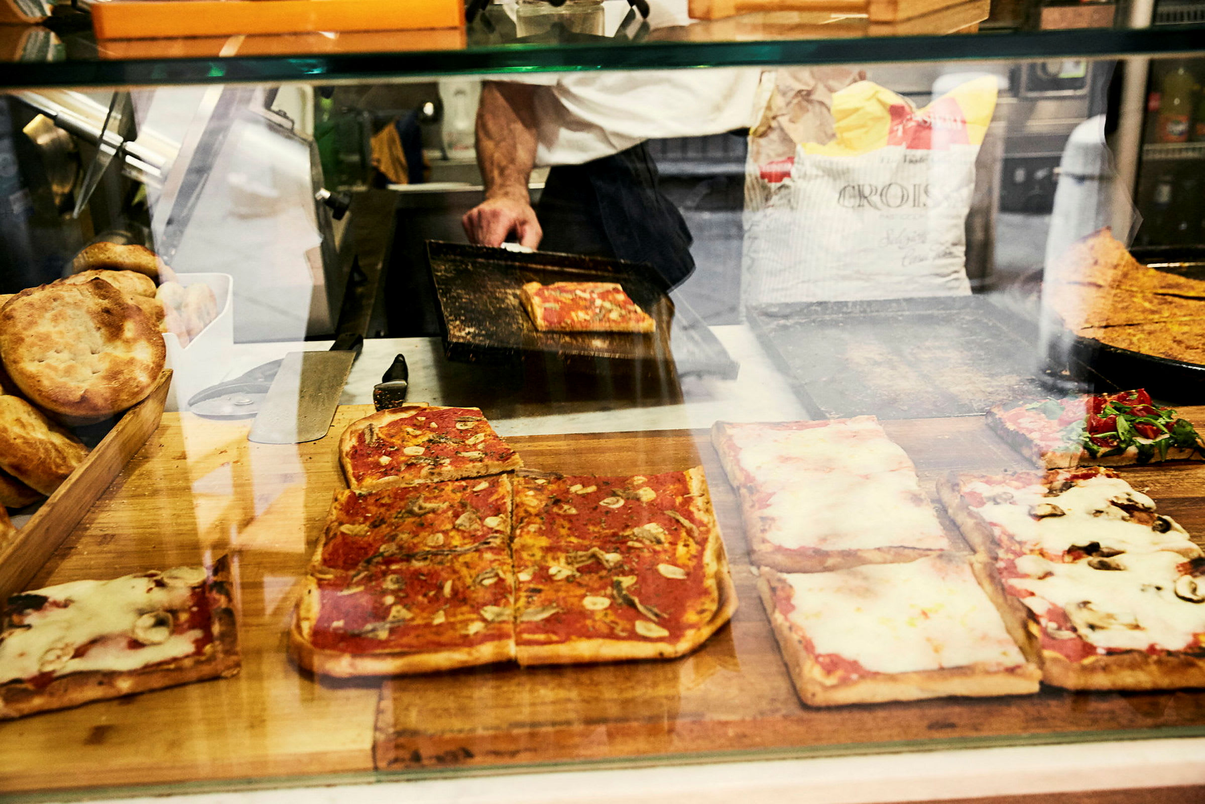 Firkanter av pizza med ulike pålegg bak en glassdisk, samt brød og en pizzaovn i bakgrunnen. (AI-generert tekst)