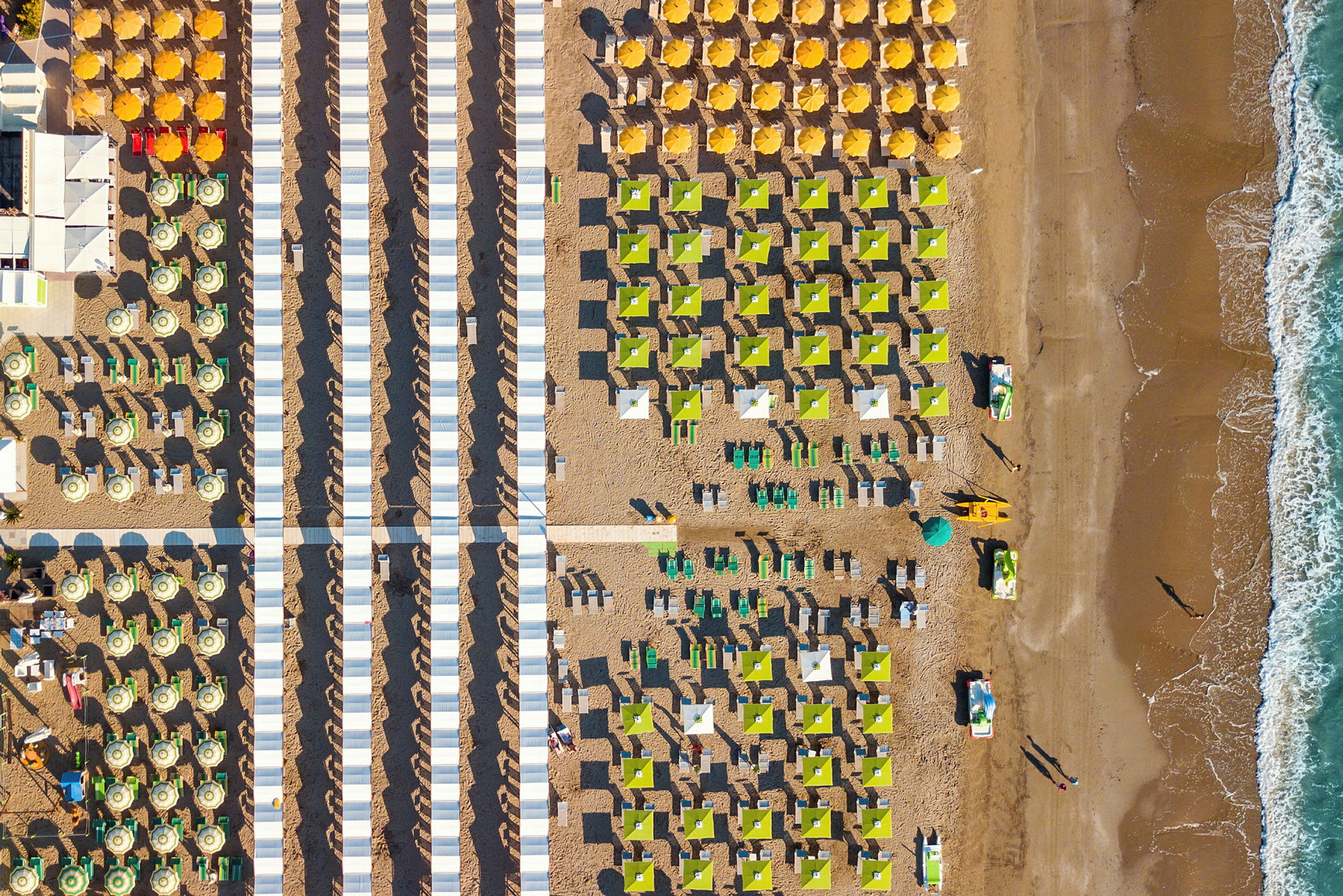 Flyfoto av en solrik strand med rekker av parasoller og solsenger arrangert i mønstre. (AI-generert tekst)