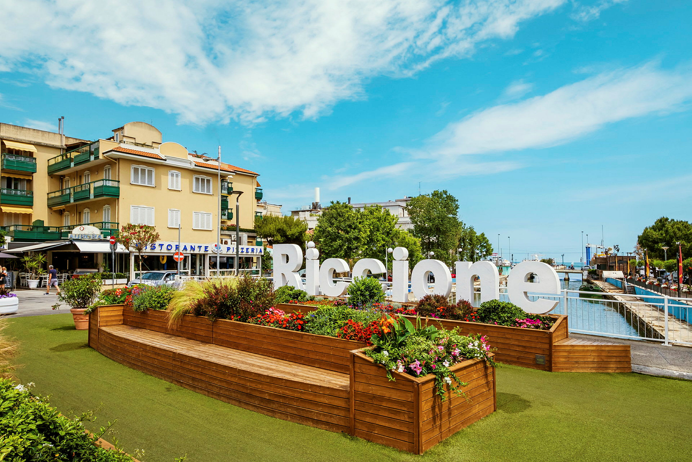 Et stort skilt med teksten "Riccione" står foran et planteområde med fargerike blomster og grønne planter. I bakgrunnen er det bygninger og en klar himmel. (AI-generert tekst)