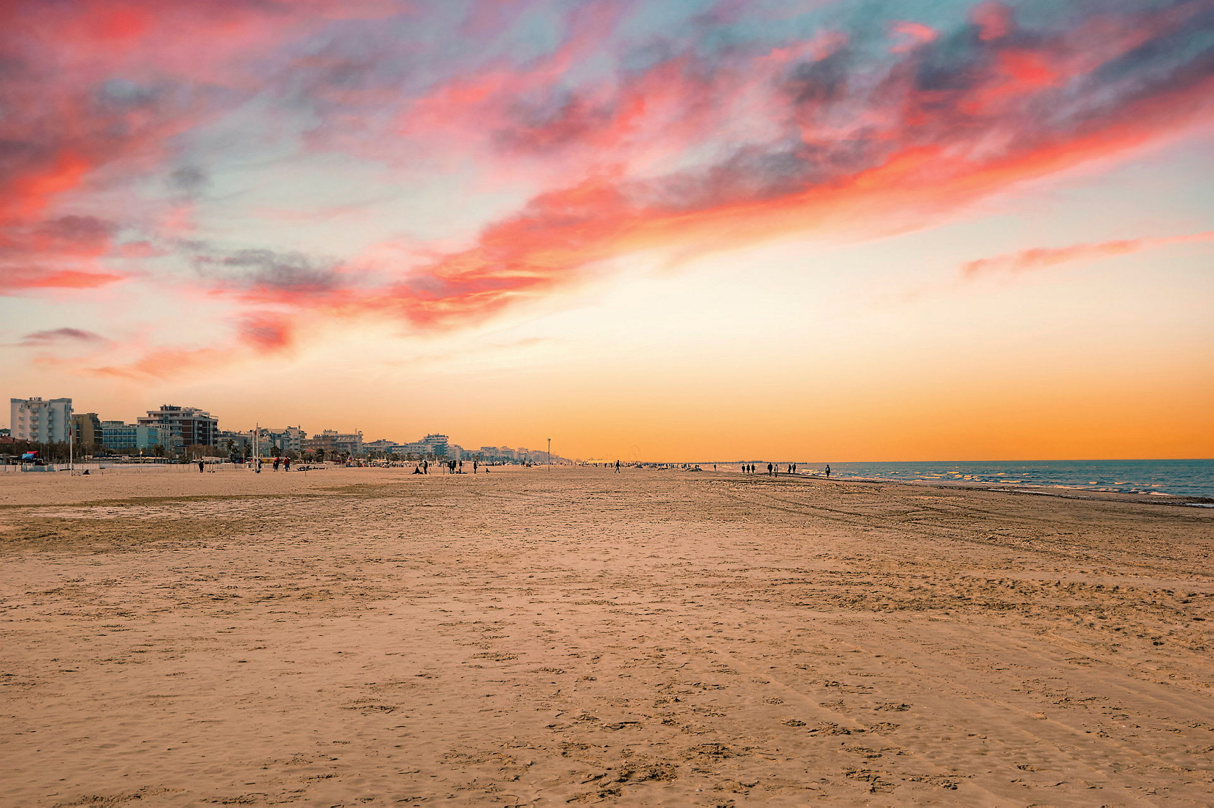 En strand ved solnedgang med en by i bakgrunnen og himmelen som lyser i oransje og rosa nyanser. (AI-generert tekst)