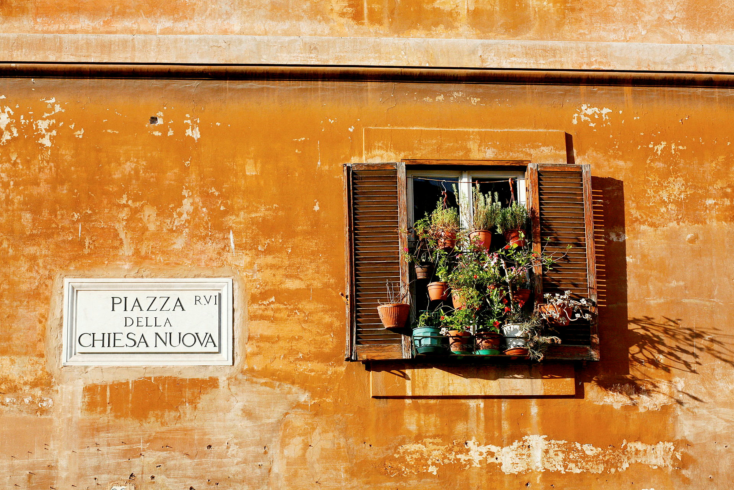 Et vindu med plantekrukker på en gul malt vegg, ved siden av et skilt med teksten 'Piazza della Chiesa Nuova'. (AI-generert tekst)