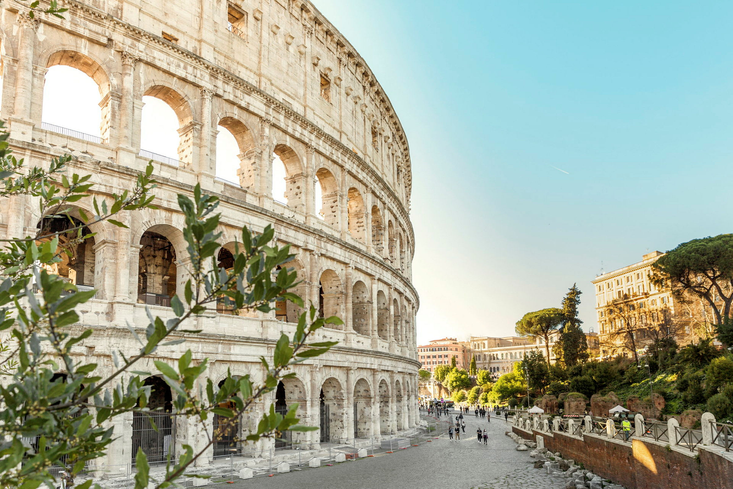 Bilde av Colosseum med omkringliggende romersk arkitektur og frodig vegetasjon under en klar blå himmel. (AI-generert tekst)