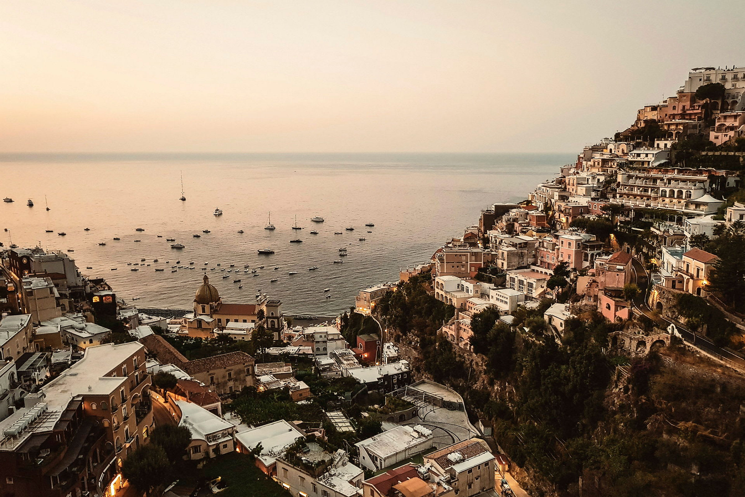 Bilde av Positano ved solnedgang med bygninger langs en skråning og havet fylt med båter. (AI-generert tekst)