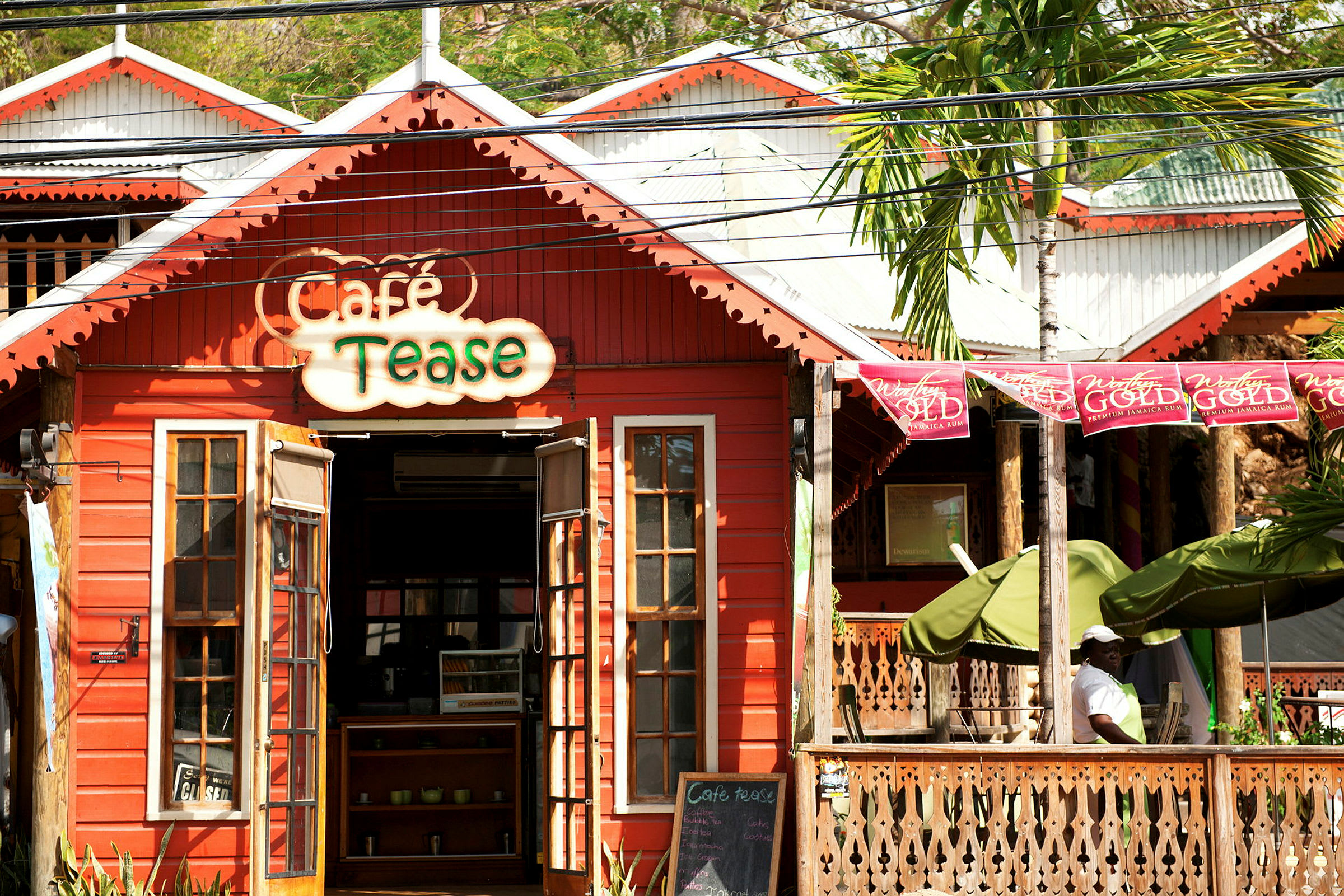 En rød trebygning med et skilt som sier "Café Tease" og åpne terrassedører, omgitt av planter og en del av en veranda. (AI-generert tekst)
