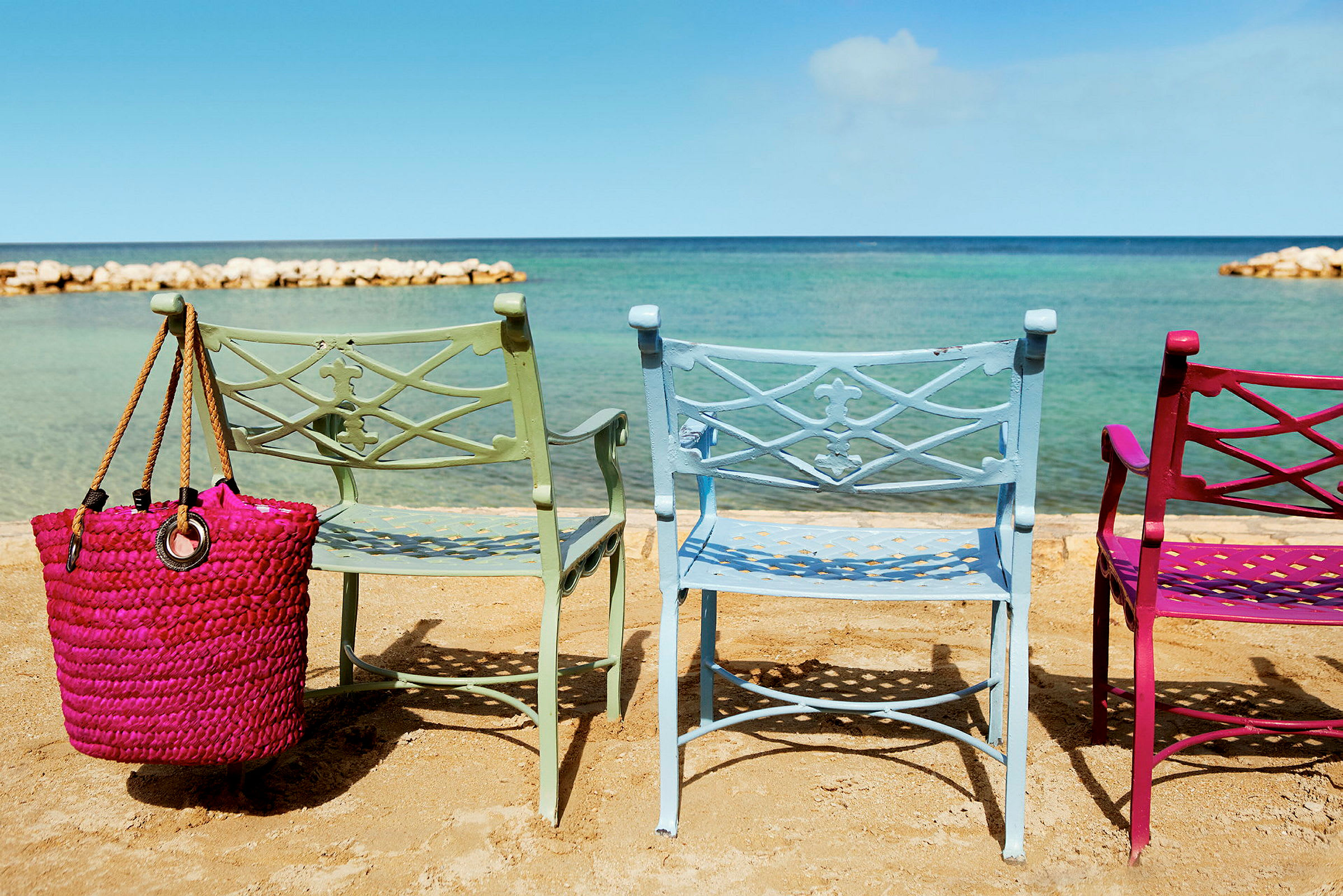 Tre fargerike stoler står på en strand med havet i bakgrunnen. En rosa strandveske henger fra en av stolene. (AI-generert tekst)