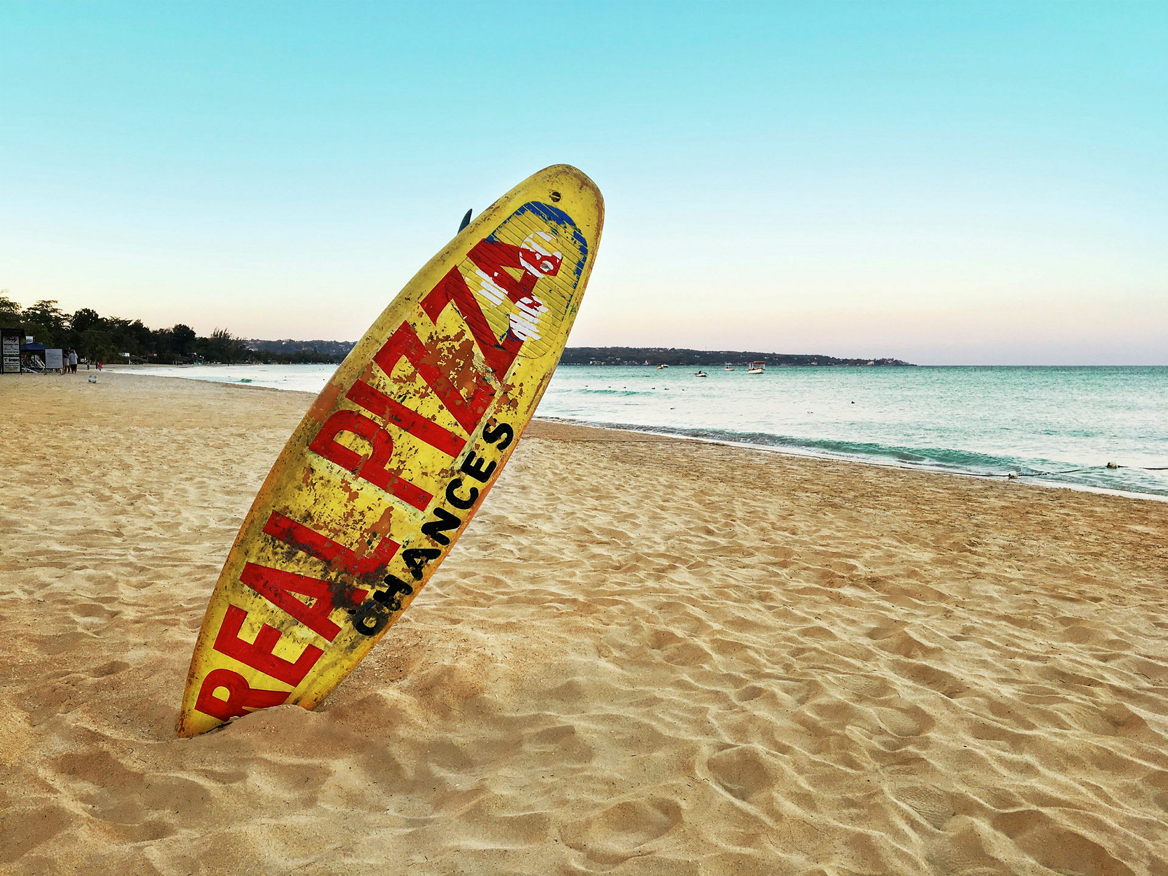 Et gul surfebrett med tekst står oppreist i sanden på en strand ved havet. (AI-generert tekst)