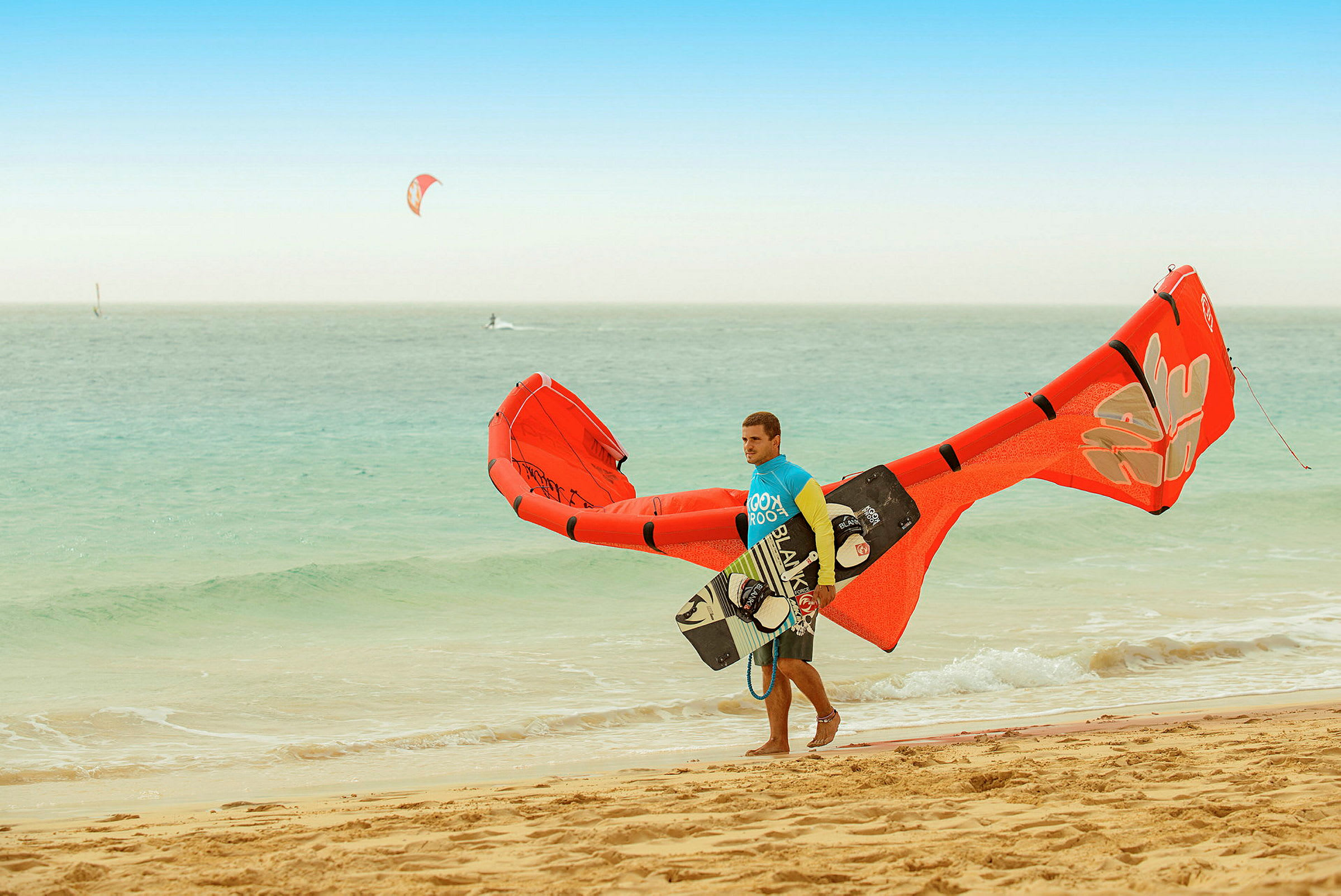 Person på en strand med kiteutstyr og surfebrett ved havet. (AI-generert tekst)