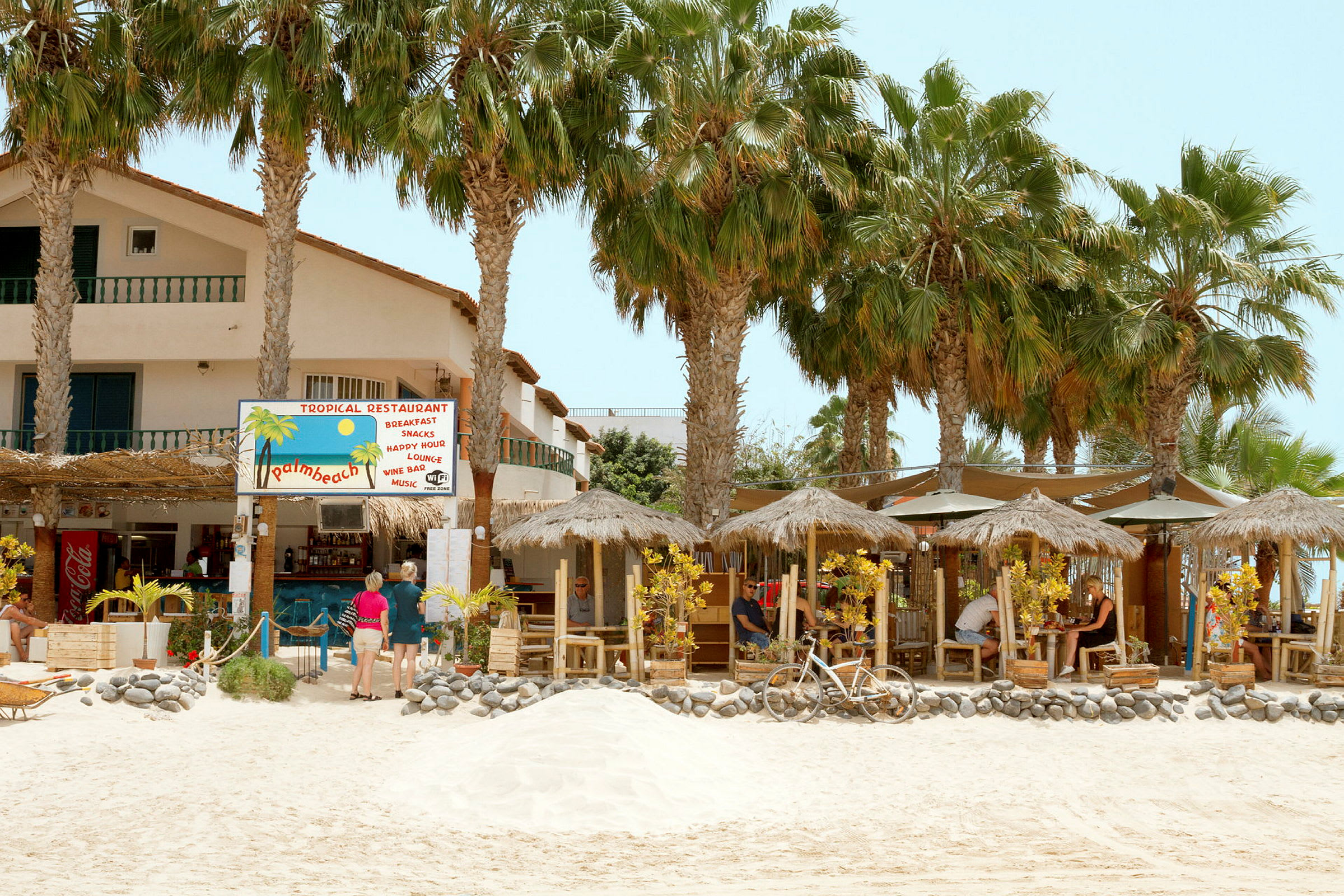 En tropisk restaurant med stråtak og palmer på en sandstrand. (AI-generert tekst)