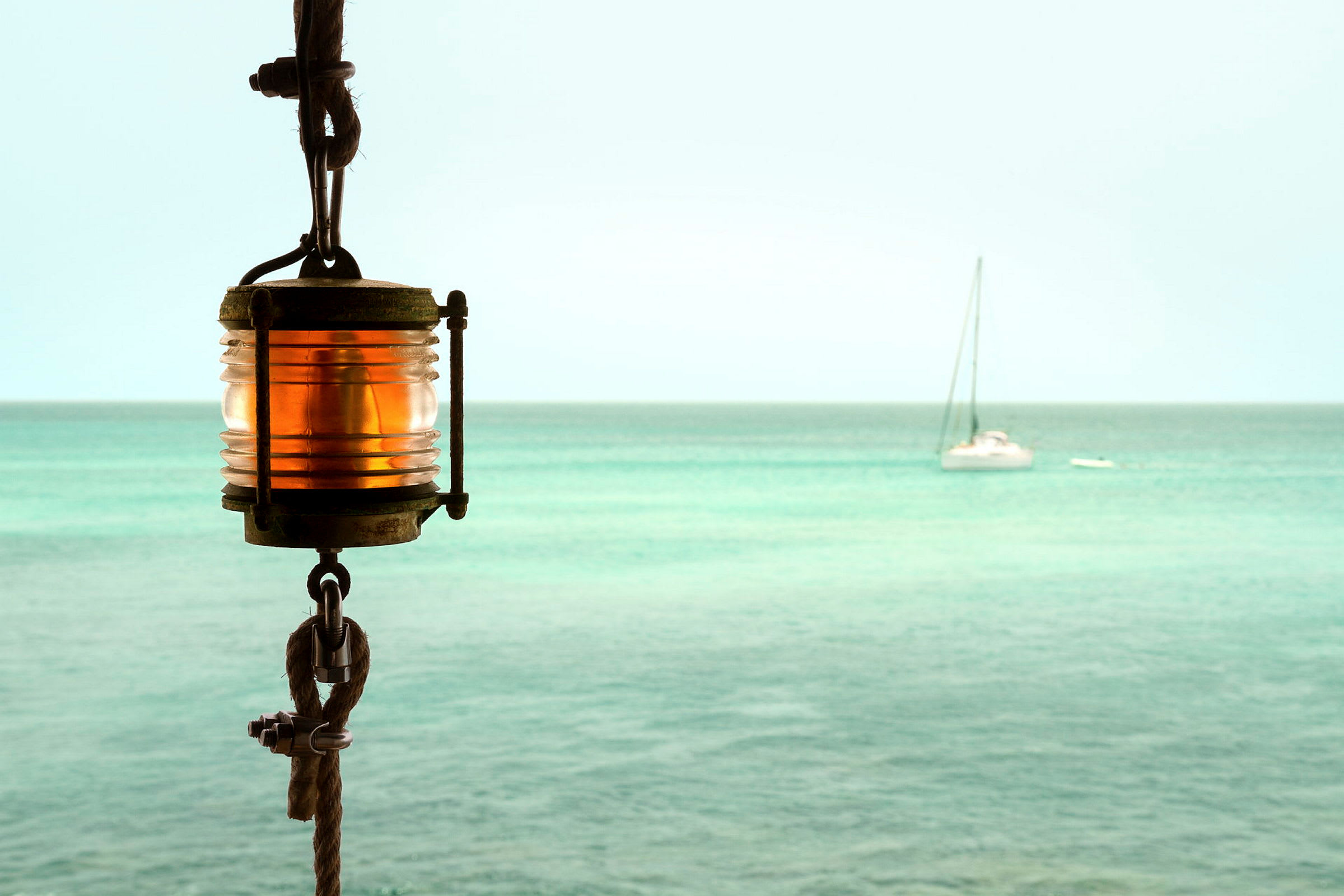 En lampe med oransje lys og tau er montert, med en seilbåt i det blå havet i bakgrunnen. (AI-generert tekst)
