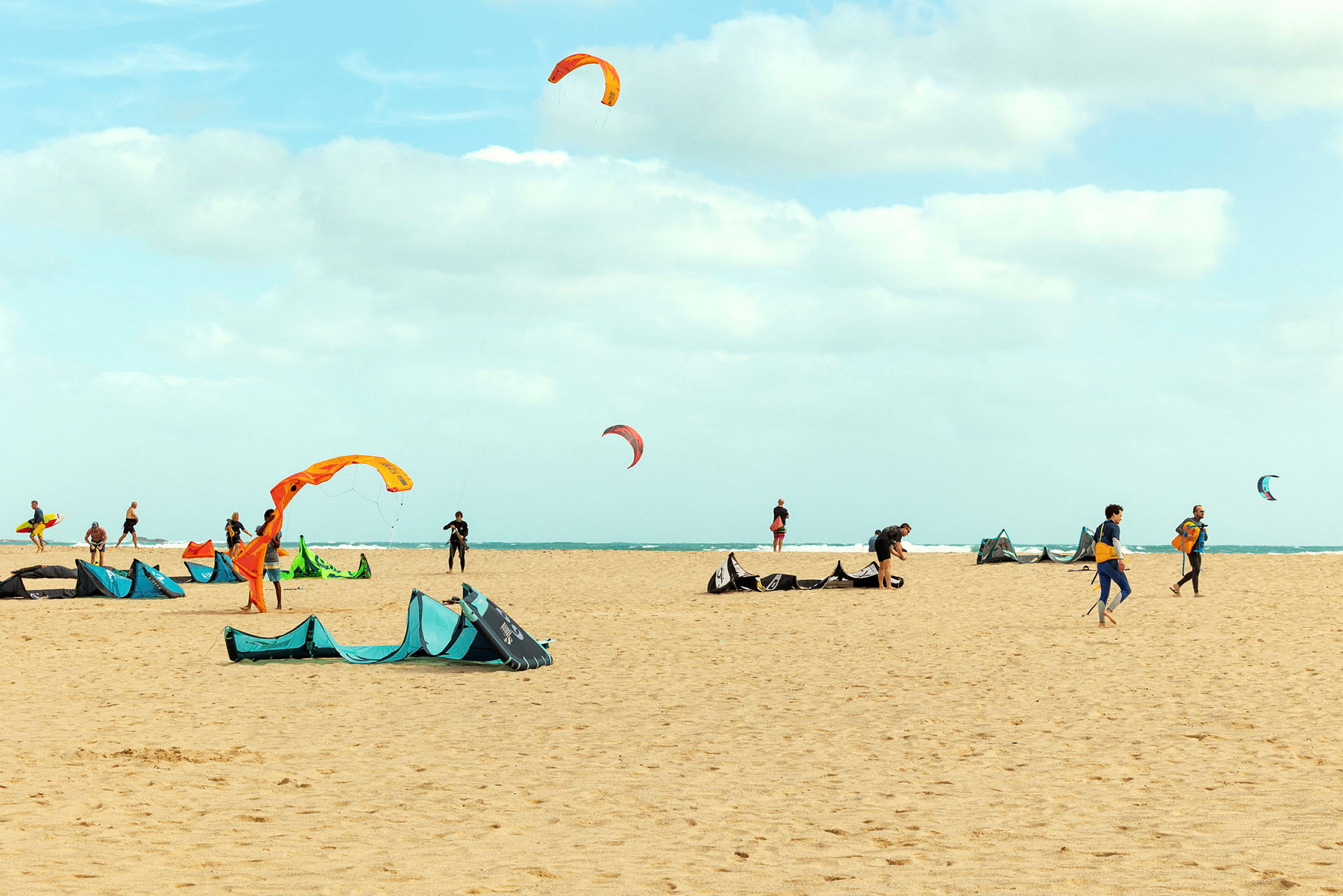Strand med personer som rigger utstyr for kitesurfing med fargerike drager på himmelen. (AI-generert tekst)