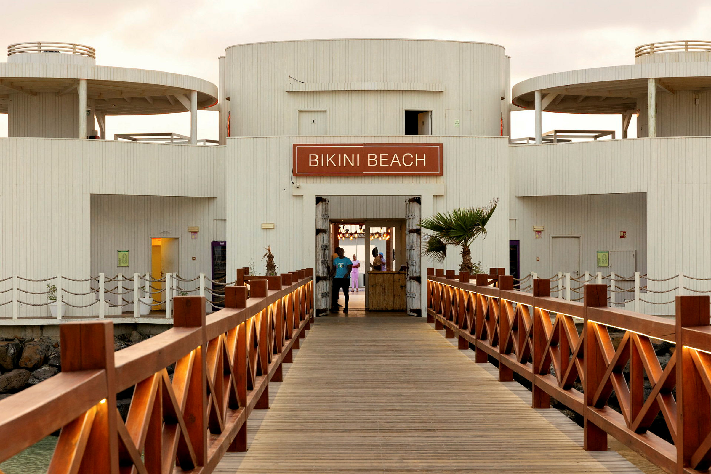 Utsikt over en trebro som leder til en bygning med skiltet 'Bikini Beach'. (AI-generert tekst)