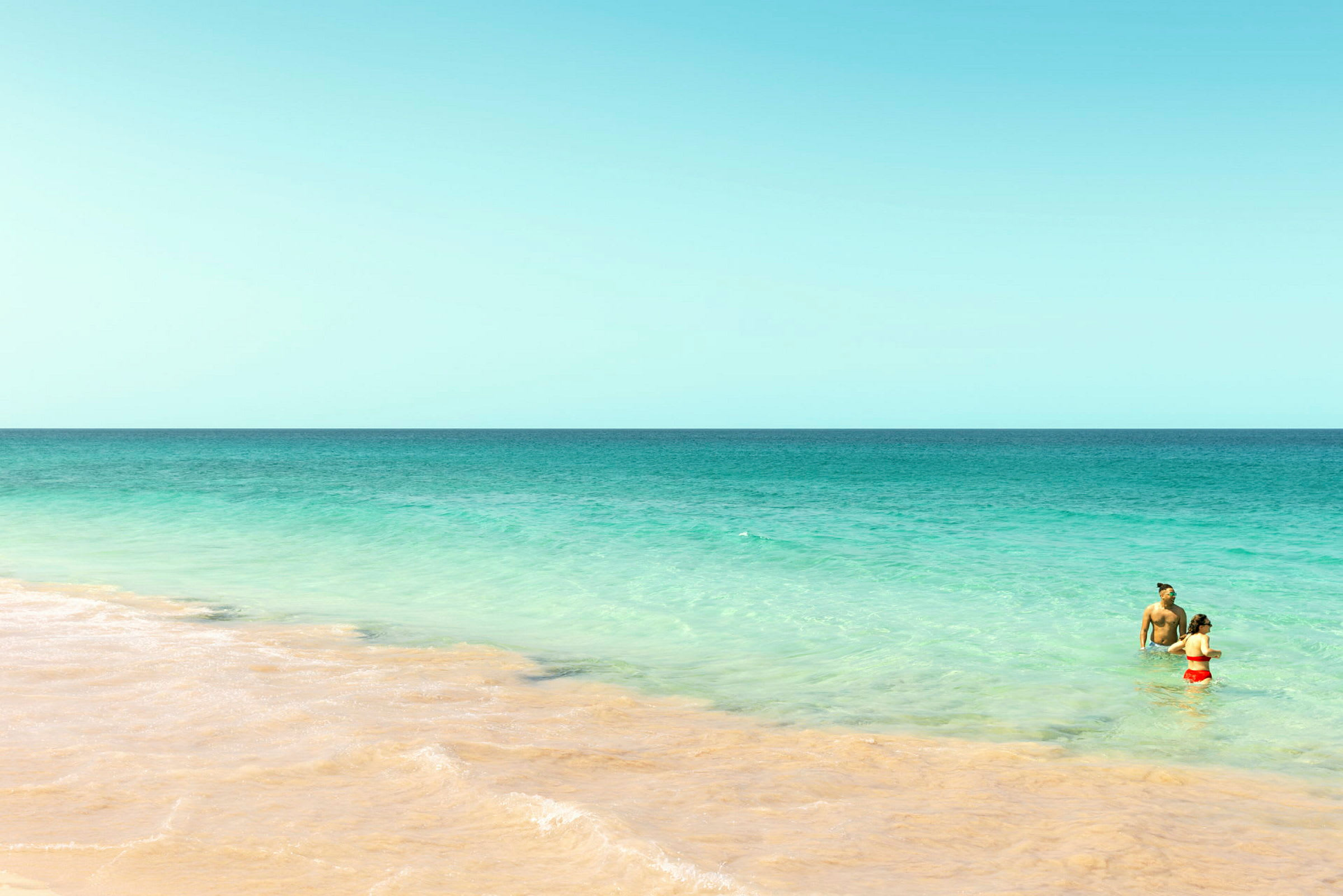 To personer står i det klare turkise havet nær en sandstrand under en skyfri himmel. (AI-generert tekst)