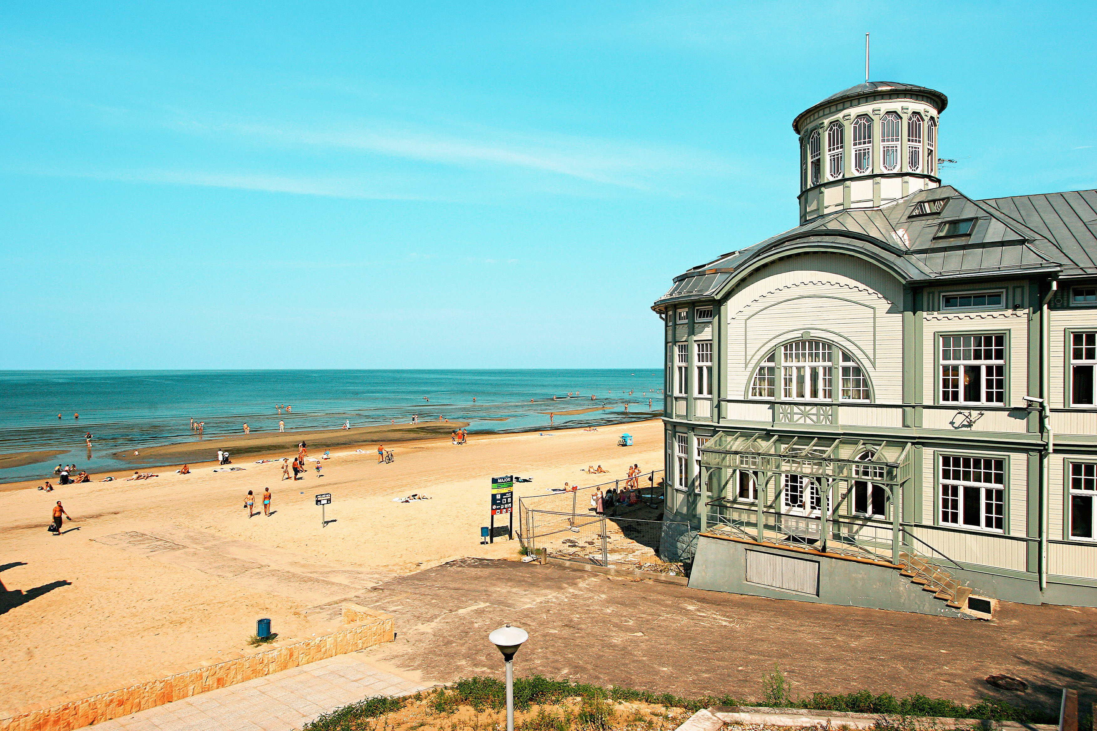 En strand med solbadende mennesker ved en rolig kystlinje, samt en større bygning med klassisk arkitektur ved stranden. (AI-generert tekst)