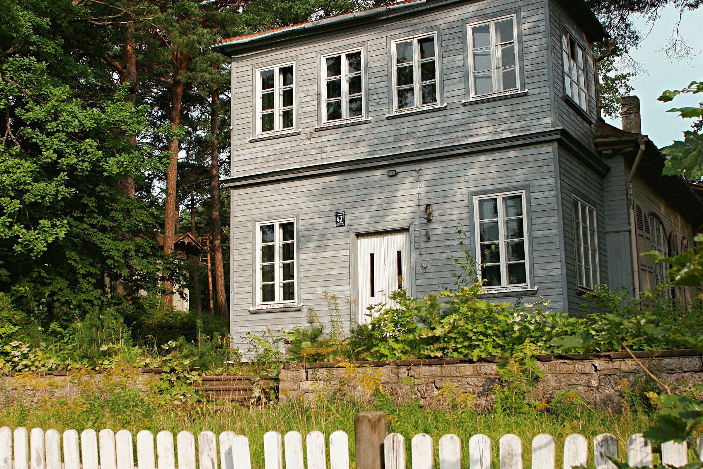 Et to-etasjes trehus malt i grå farge, omgitt av vegetasjon og med et hvitt gjerde i forgrunnen. (AI-generert tekst)